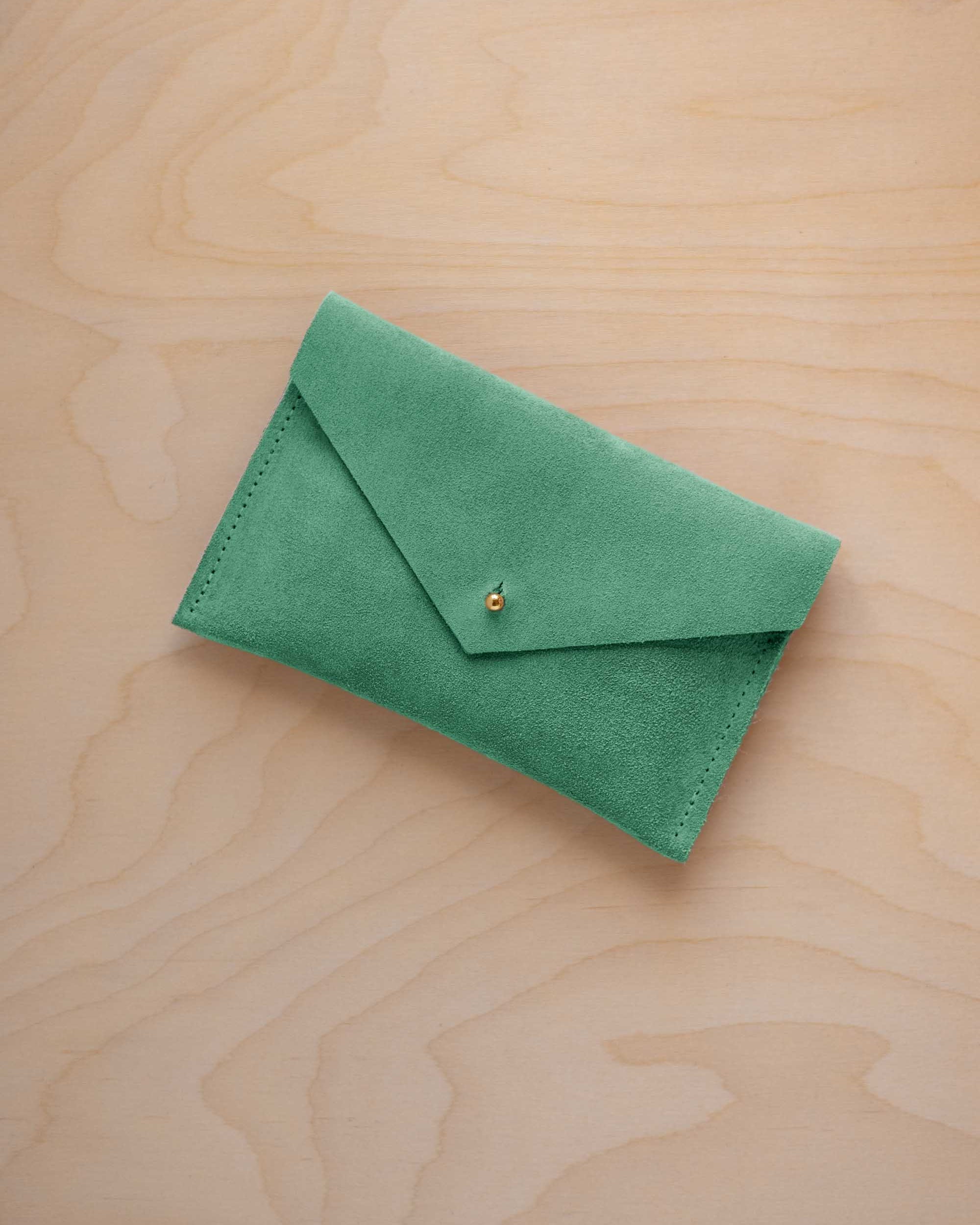 A small seafoam green coloured suede envelope-style pouch with a gold button, resting on a light wood grain surface.