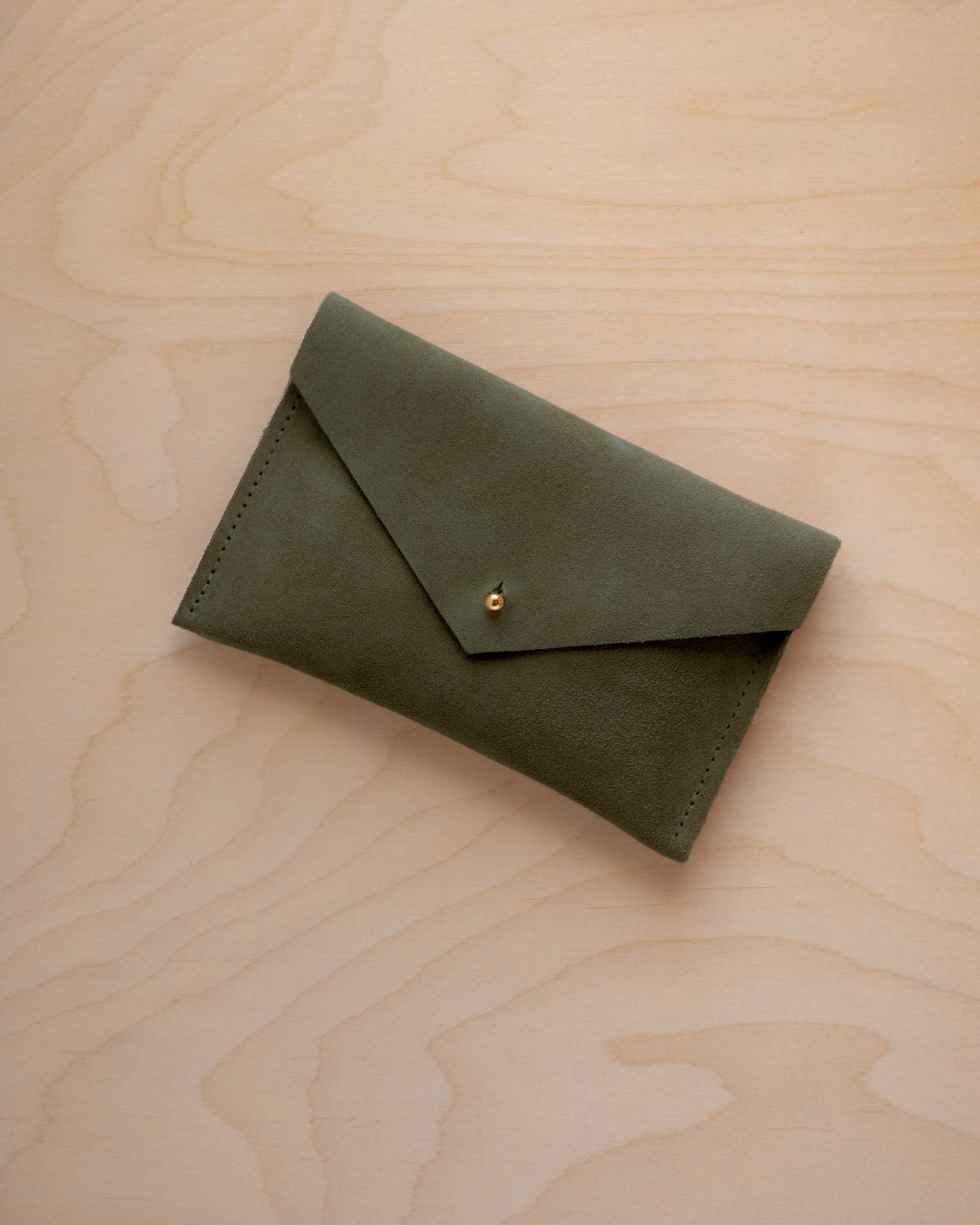 A small soft olive green coloured suede envelope-style pouch with a gold button, resting on a light wood grain surface.