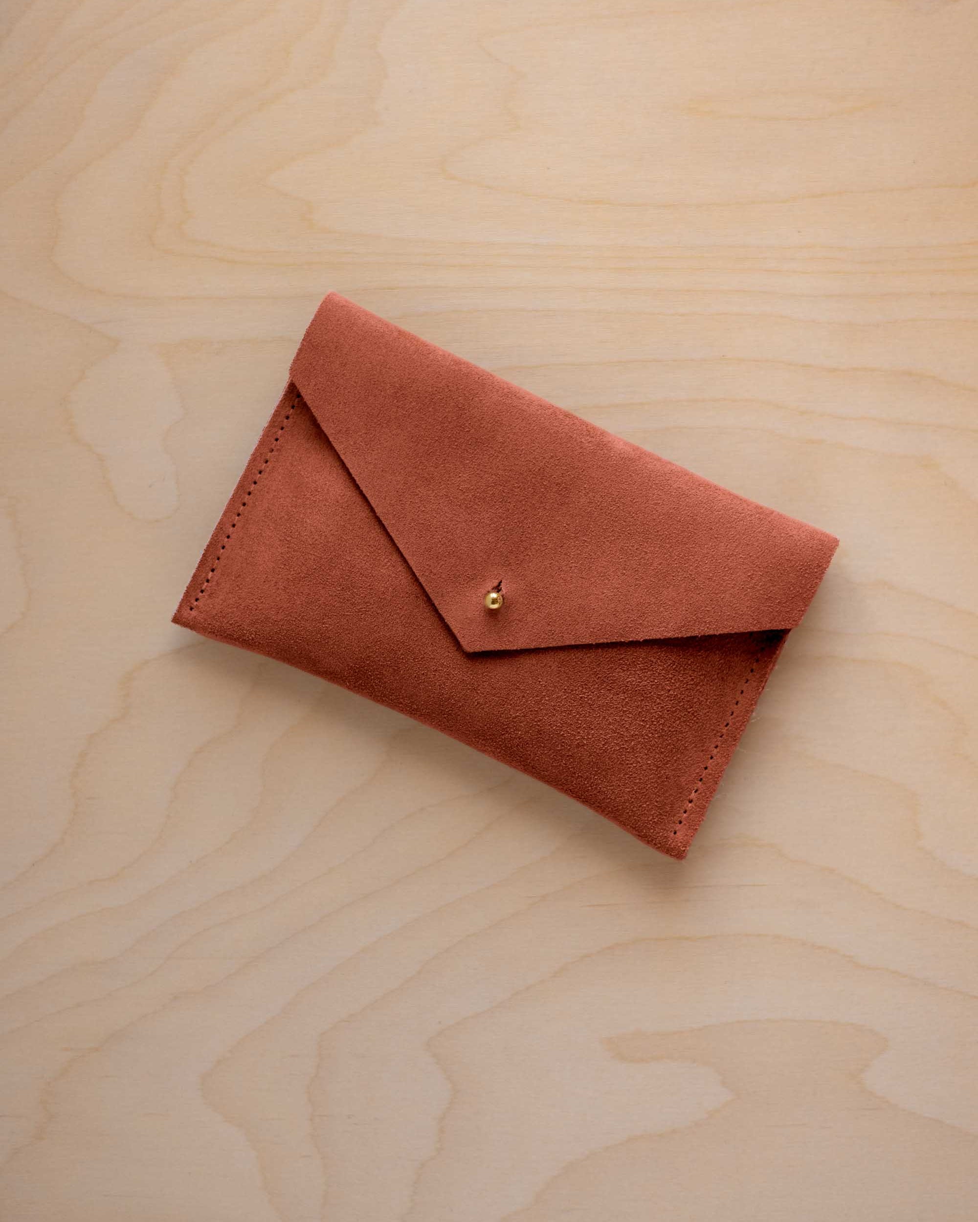 A small terracotta coloured suede envelope-style pouch with a gold button, resting on a light wood grain surface.