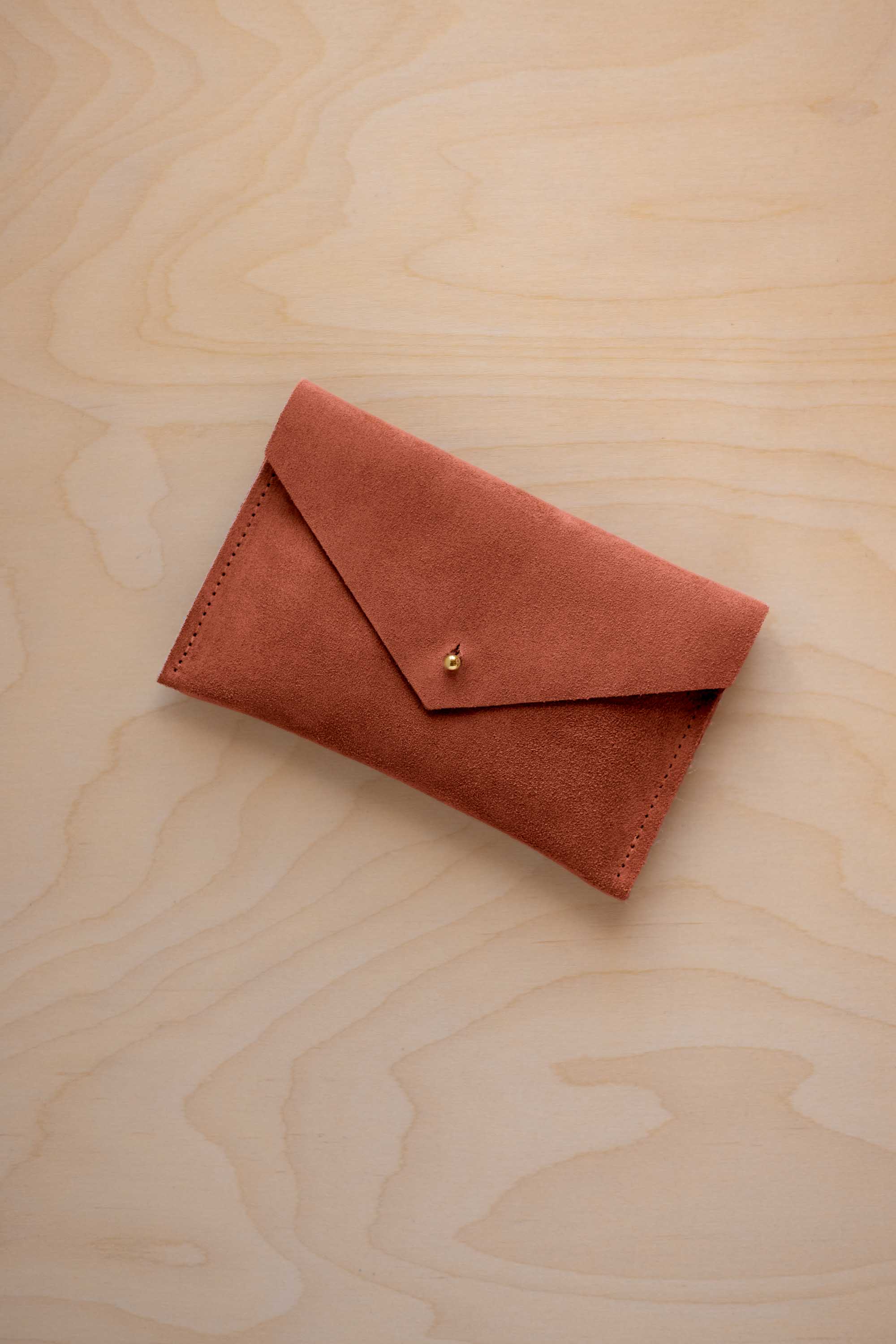 A small terracotta coloured suede envelope-style pouch with a gold button, resting on a light wood grain surface.