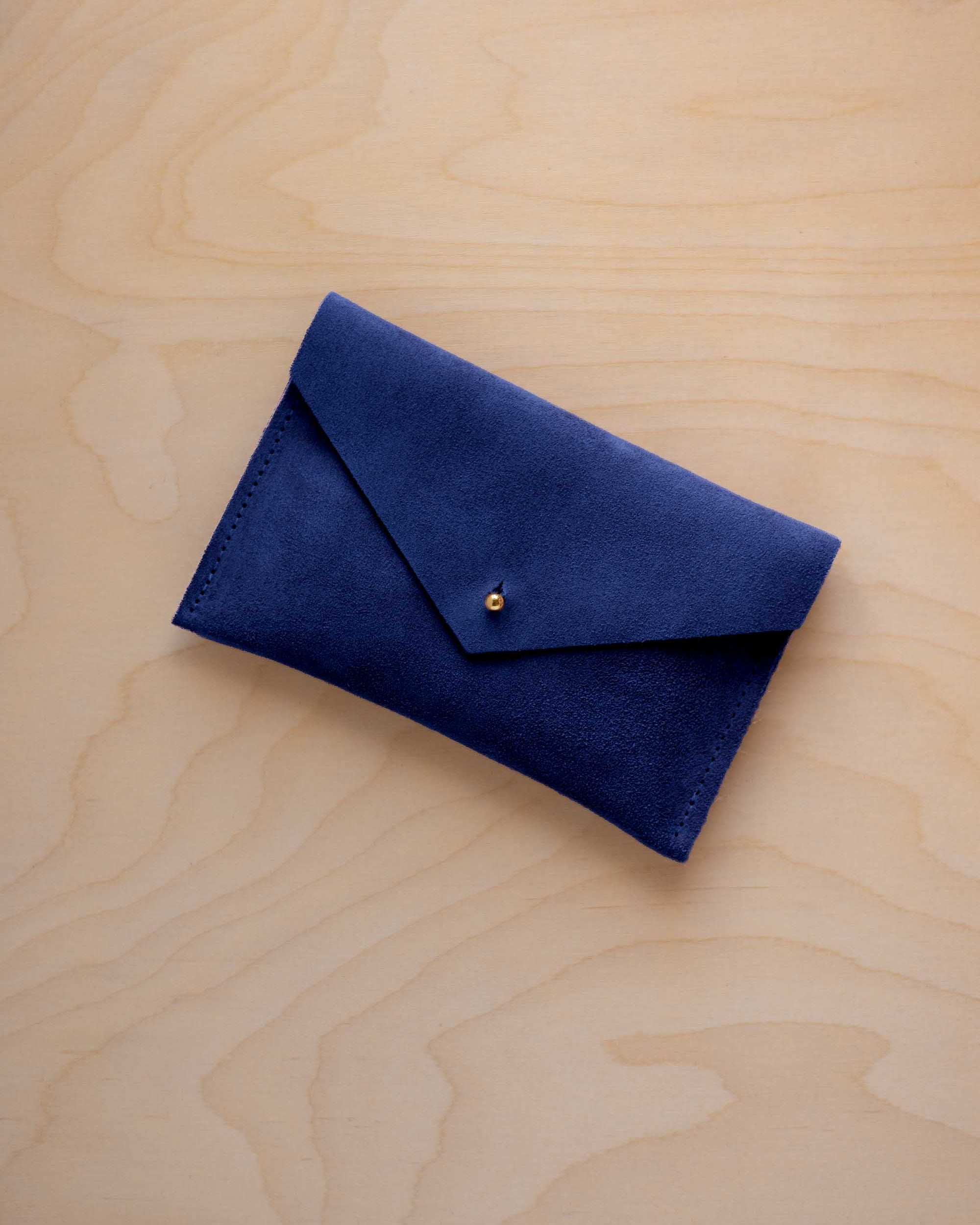 A small ultramarine blue coloured suede envelope-style pouch with a gold button, resting on a light wood grain surface.