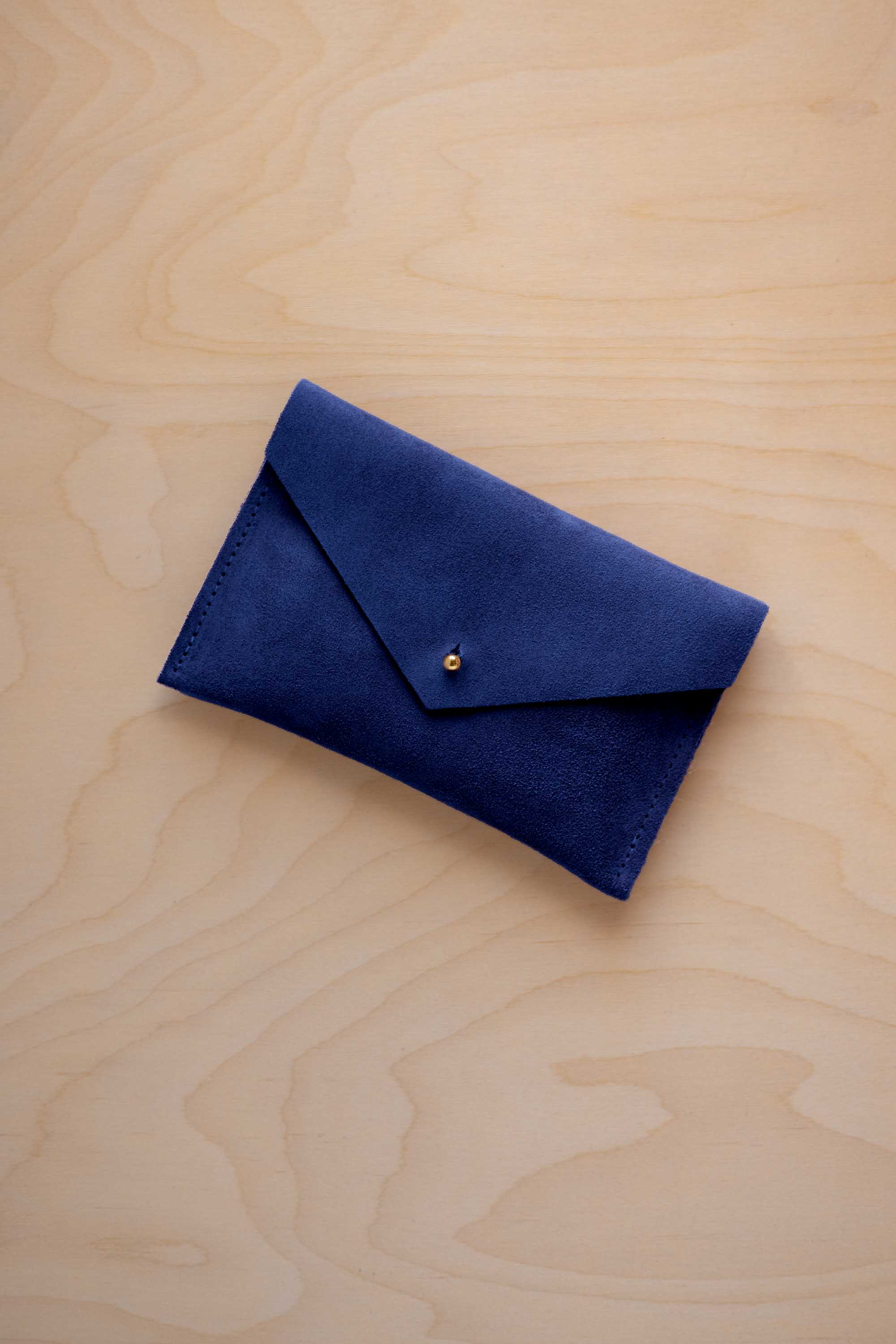 A small ultramarine blue coloured suede envelope-style pouch with a gold button, resting on a light wood grain surface.