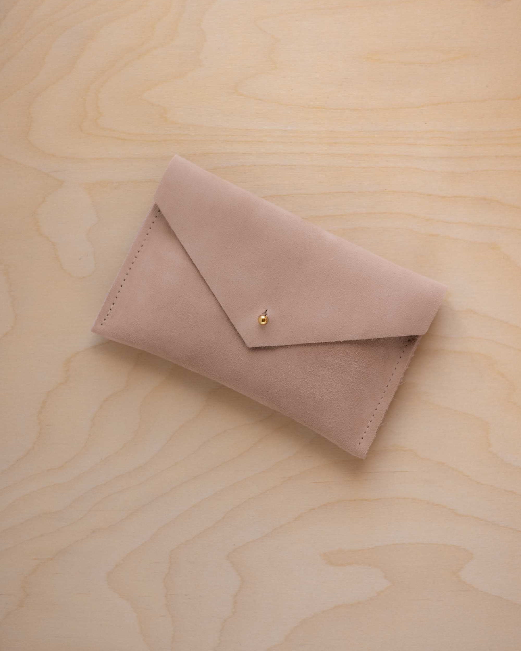 A small warm sand coloured suede envelope-style pouch with a gold button, resting on a light wood grain surface.