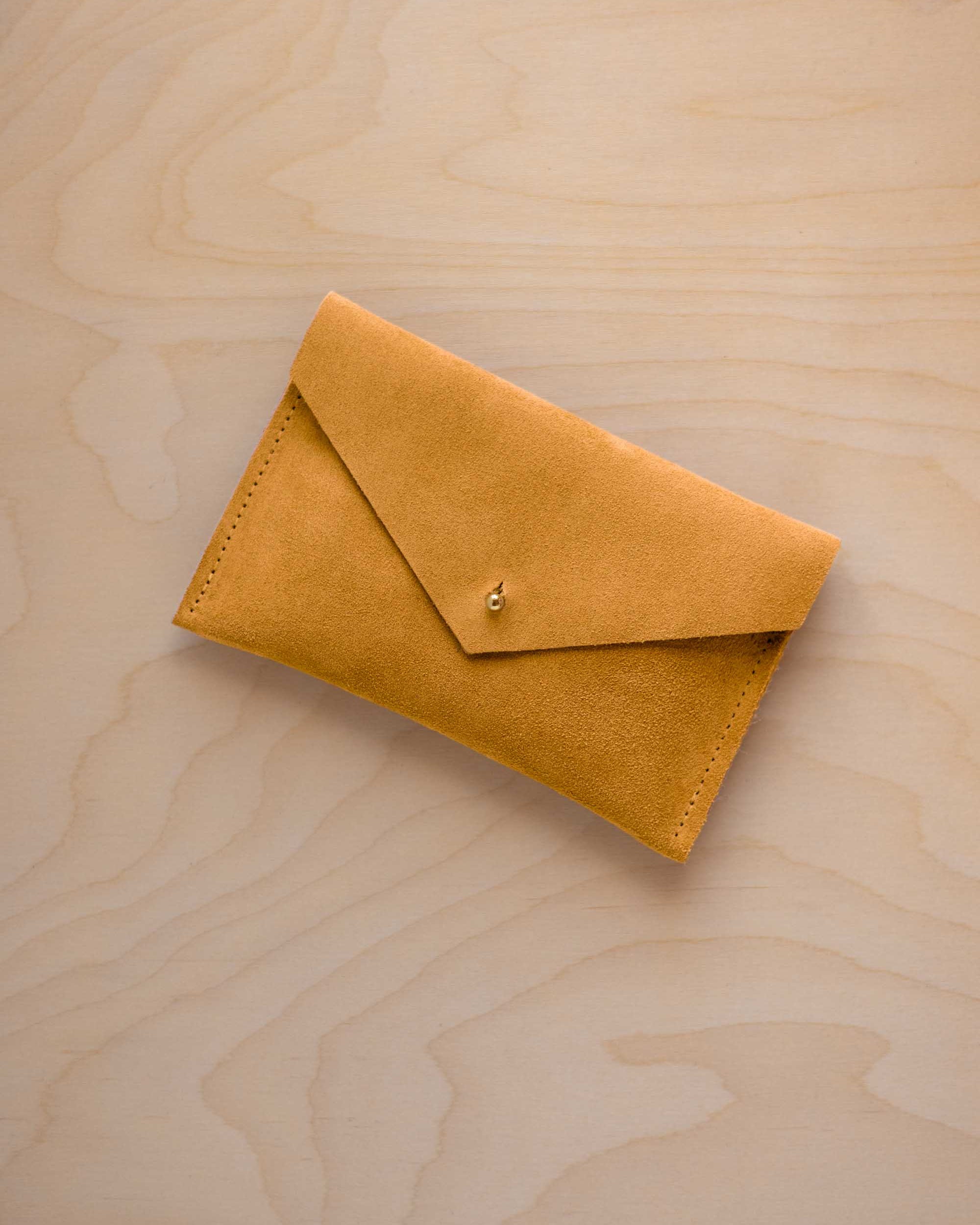 A small golden ochre coloured suede envelope-style pouch with a gold button, resting on a light wood grain surface.