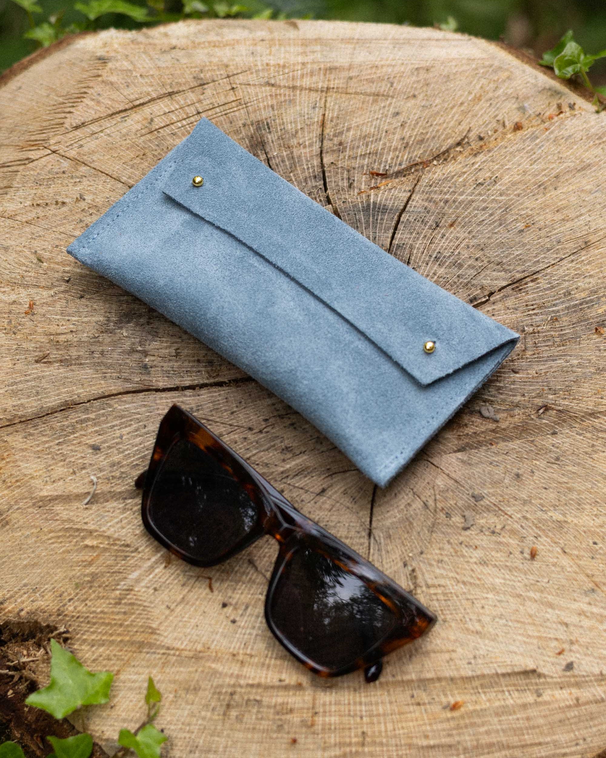 A light blue suede eyeglasses case with gold-tone buttons, shown on a rustic tree stump outdoors with tortoiseshell sunglasses resting in front.