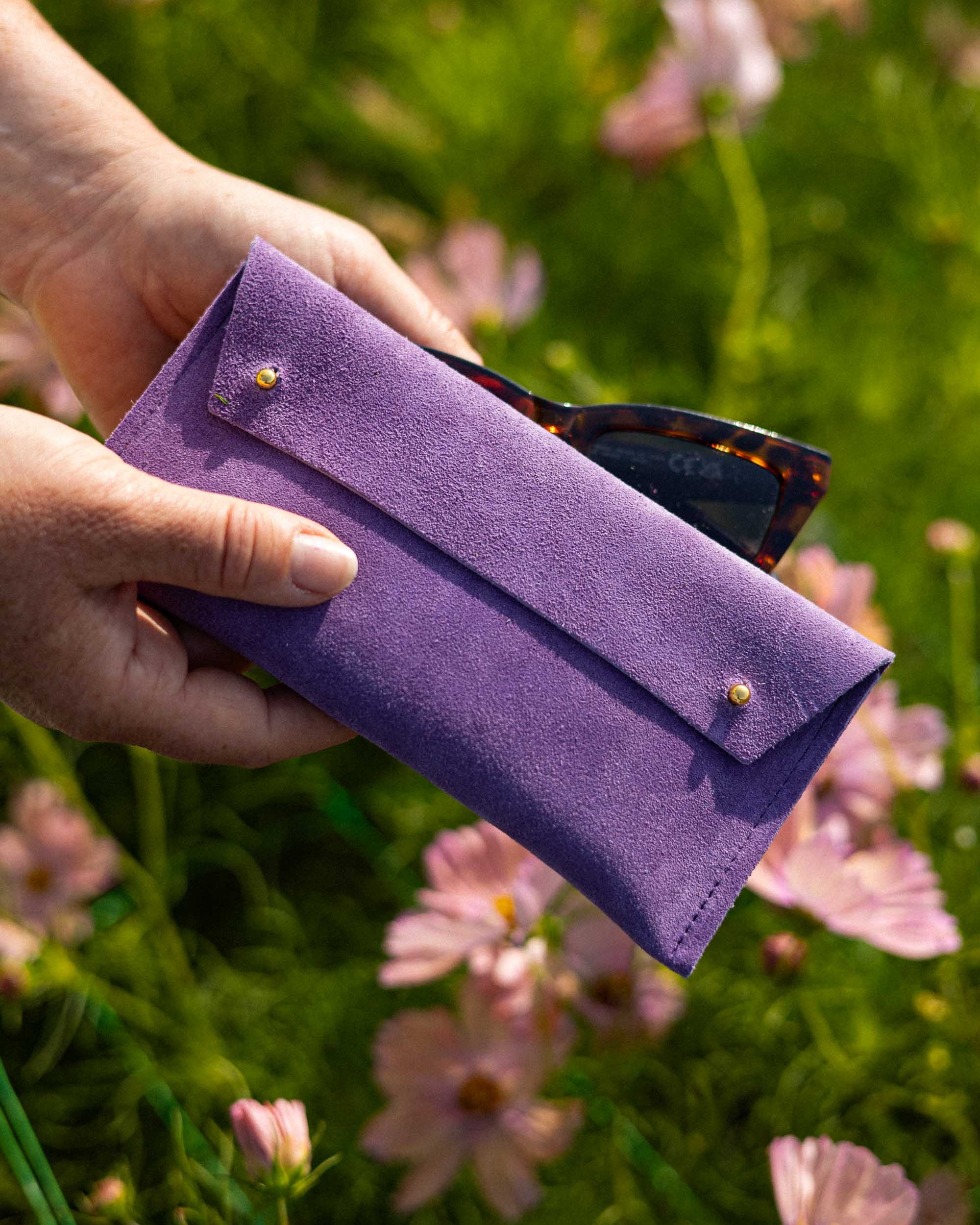 A person's hands holding a purple suede sunglasses case with gold buttons, with tortoiseshell sunglasses partially tucked inside, set against a blurred background of green grass and pink flowers.