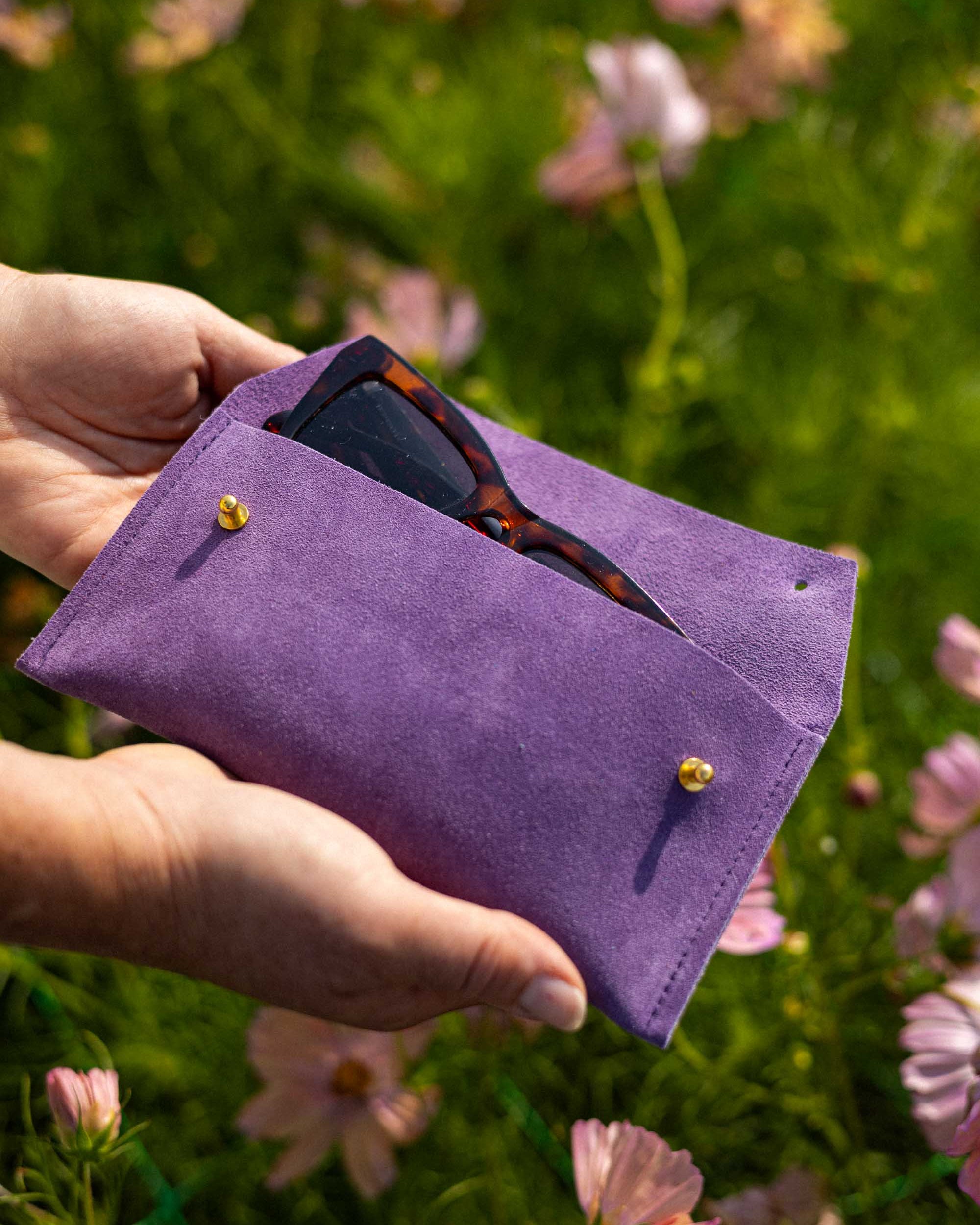 A person's hands holding a purple suede sunglasses pouch with gold buttons, containing tortoiseshell sunglasses, set against a blurred background of green grass and pink flowers.