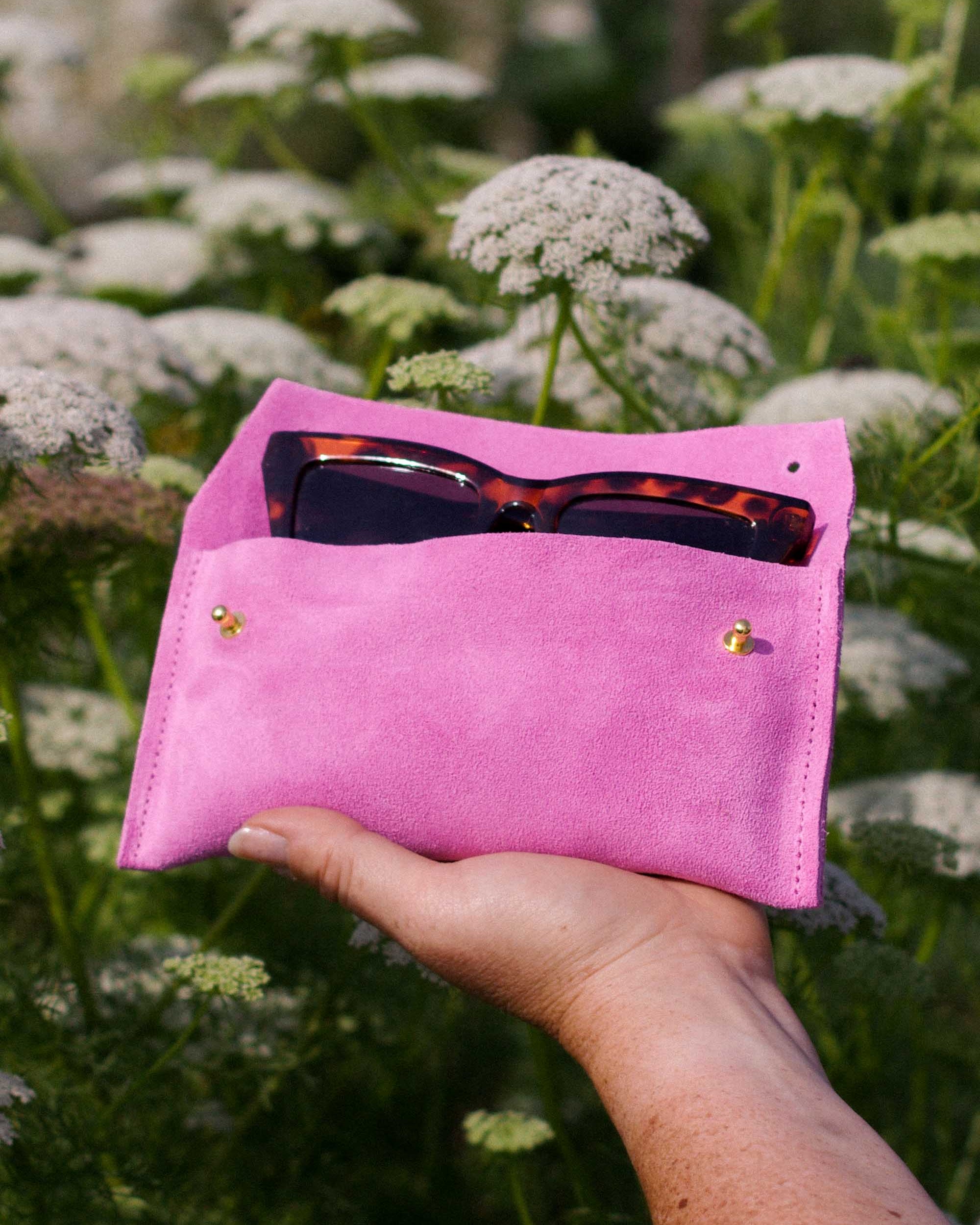 A hand holding a bright pink suede pouch or eyeglasses holder outdoors among white flowers. Tortoiseshell sunglasses are tucked into the top pocket.