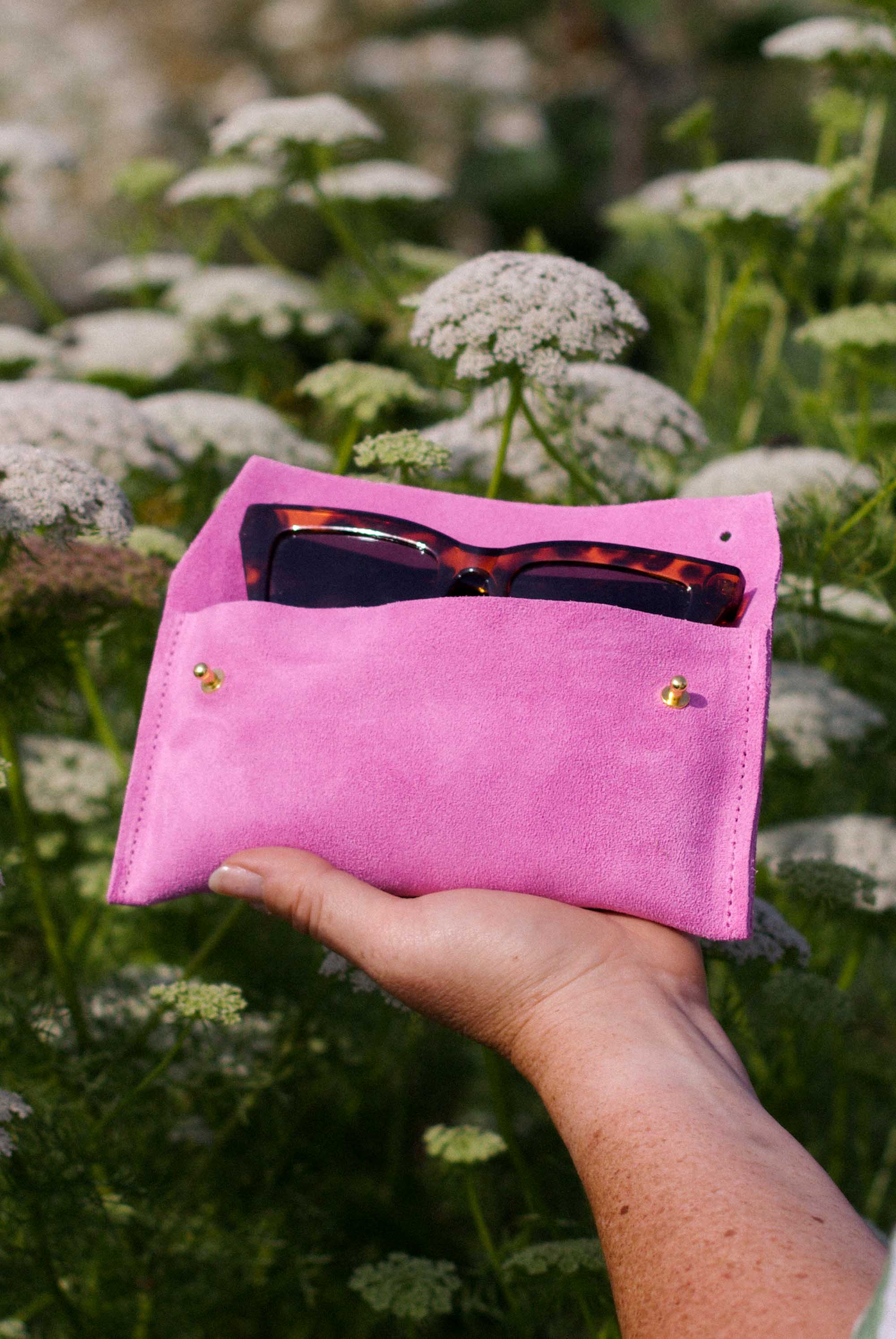 A hand holding a bright pink suede pouch or eyeglasses holder outdoors among white flowers. Tortoiseshell sunglasses are tucked into the top pocket.