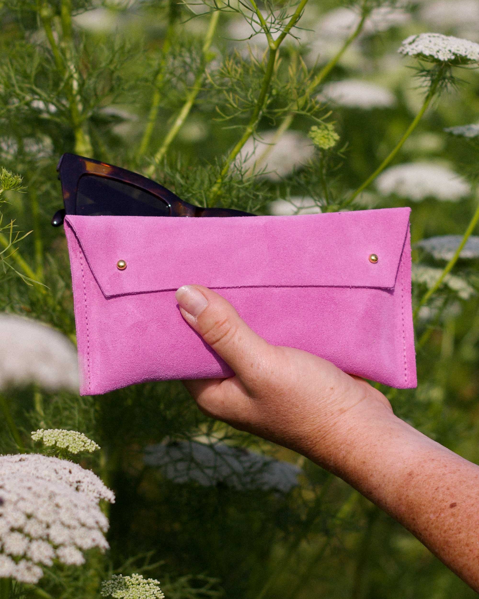 A hand holding a bright pink sunglasses case with gold-tone buttons, set against a blurred background of green plants and white flowers.