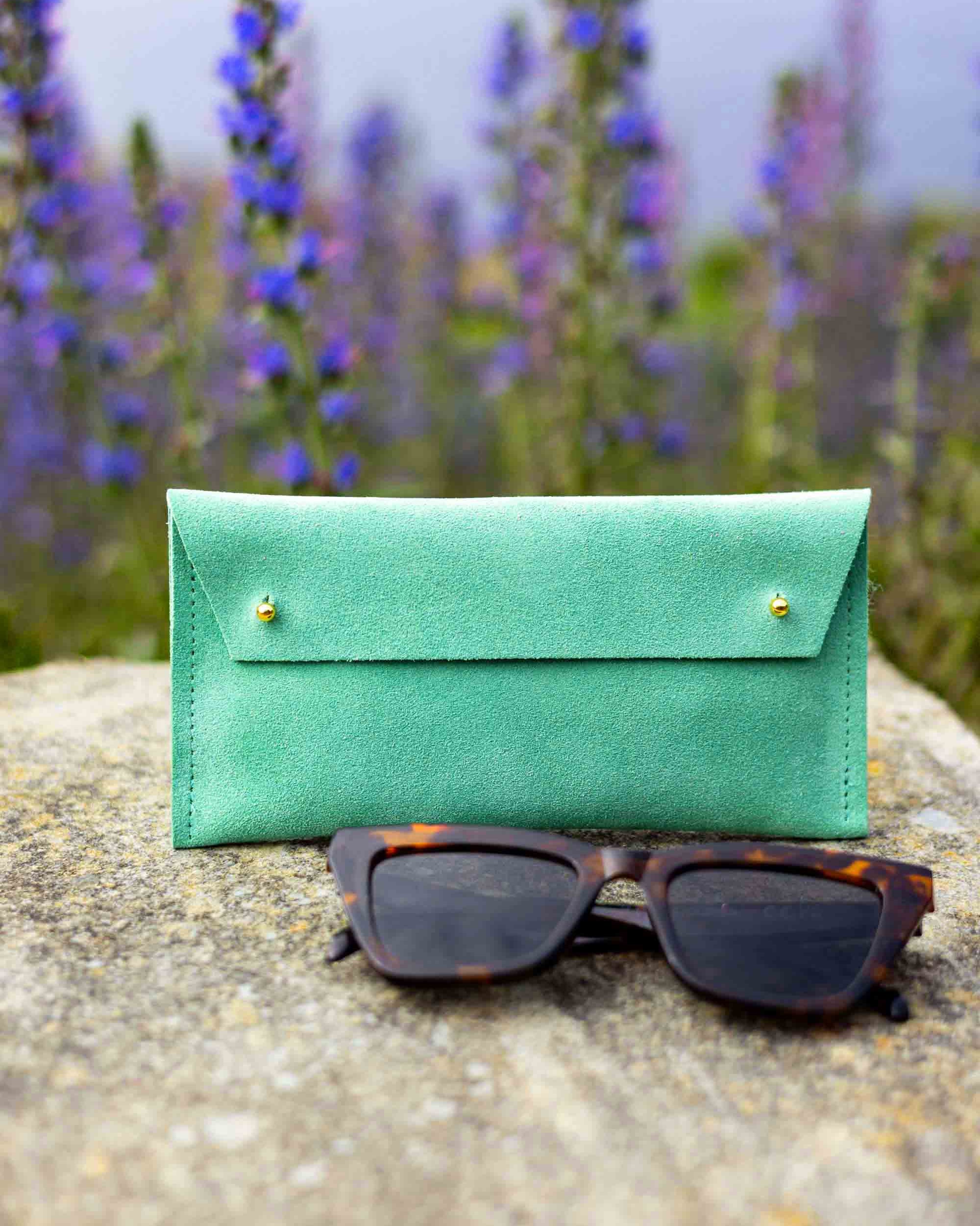 A soft green leather sunglasses case with gold-tone buttons stands on a stone surface outdoors, with tortoiseshell sunglasses placed in front and blurred purple flowers in the background.