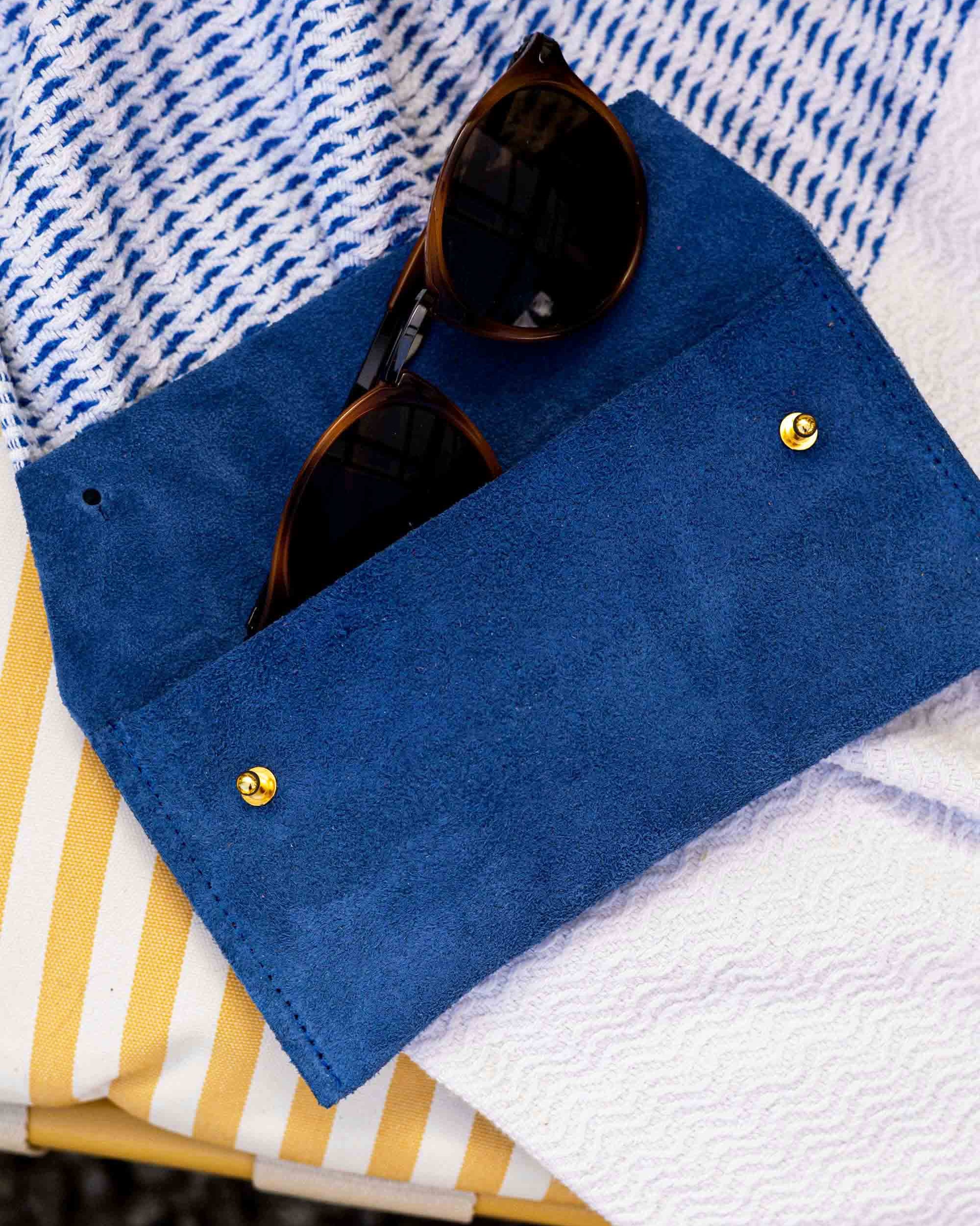 A royal blue blue suede sunglasses case with gold snap buttons, holding a pair of sunglasses, styled on a blue and white striped textile over a yellow and white striped surface.