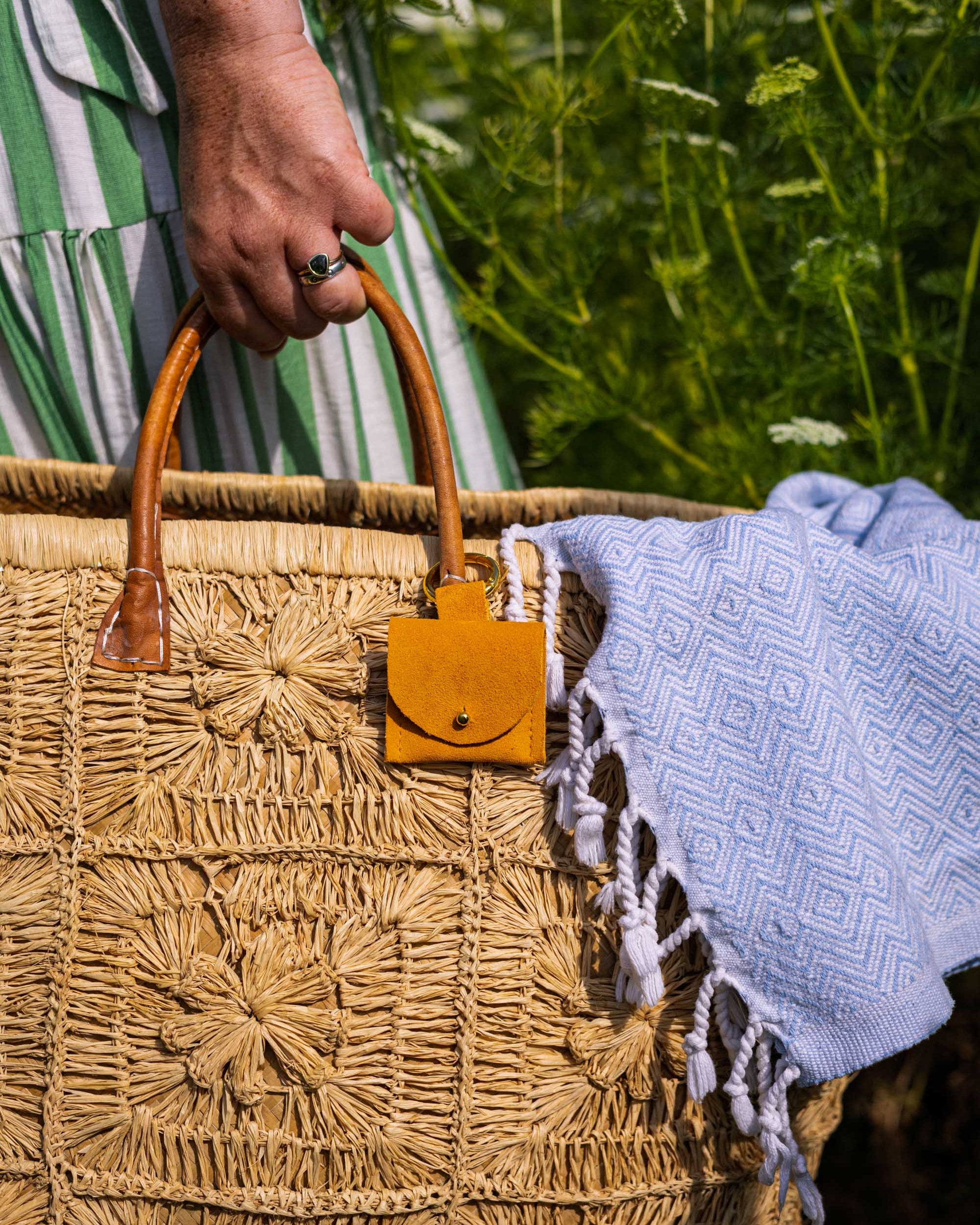 A close-up of a woven straw tote bag with a small mustard yellow suede pouch with gold clip hanging from a tan leather handle, held by a hand outdoors with a white fringed blanket draped over the side.