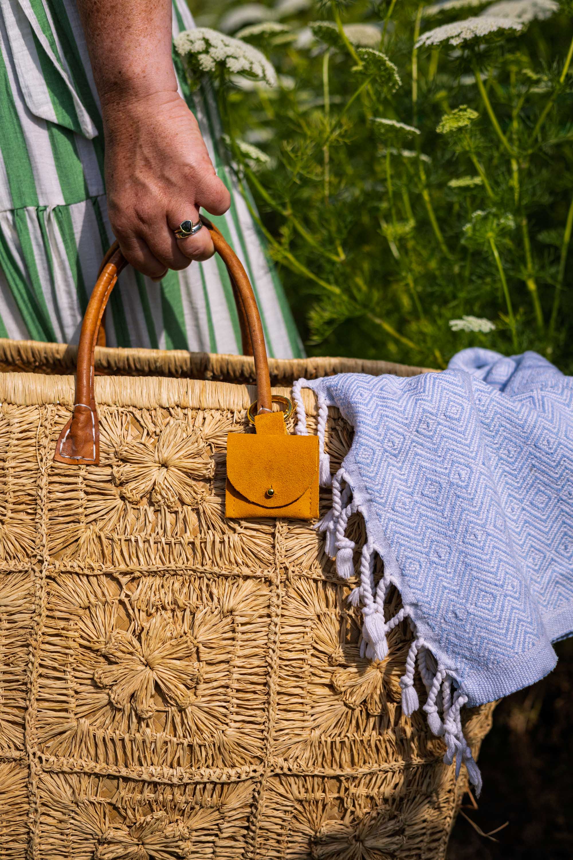 A close-up of a woven straw tote bag with a small mustard yellow suede pouch with gold clip hanging from a tan leather handle, held by a hand outdoors with a white fringed blanket draped over the side.