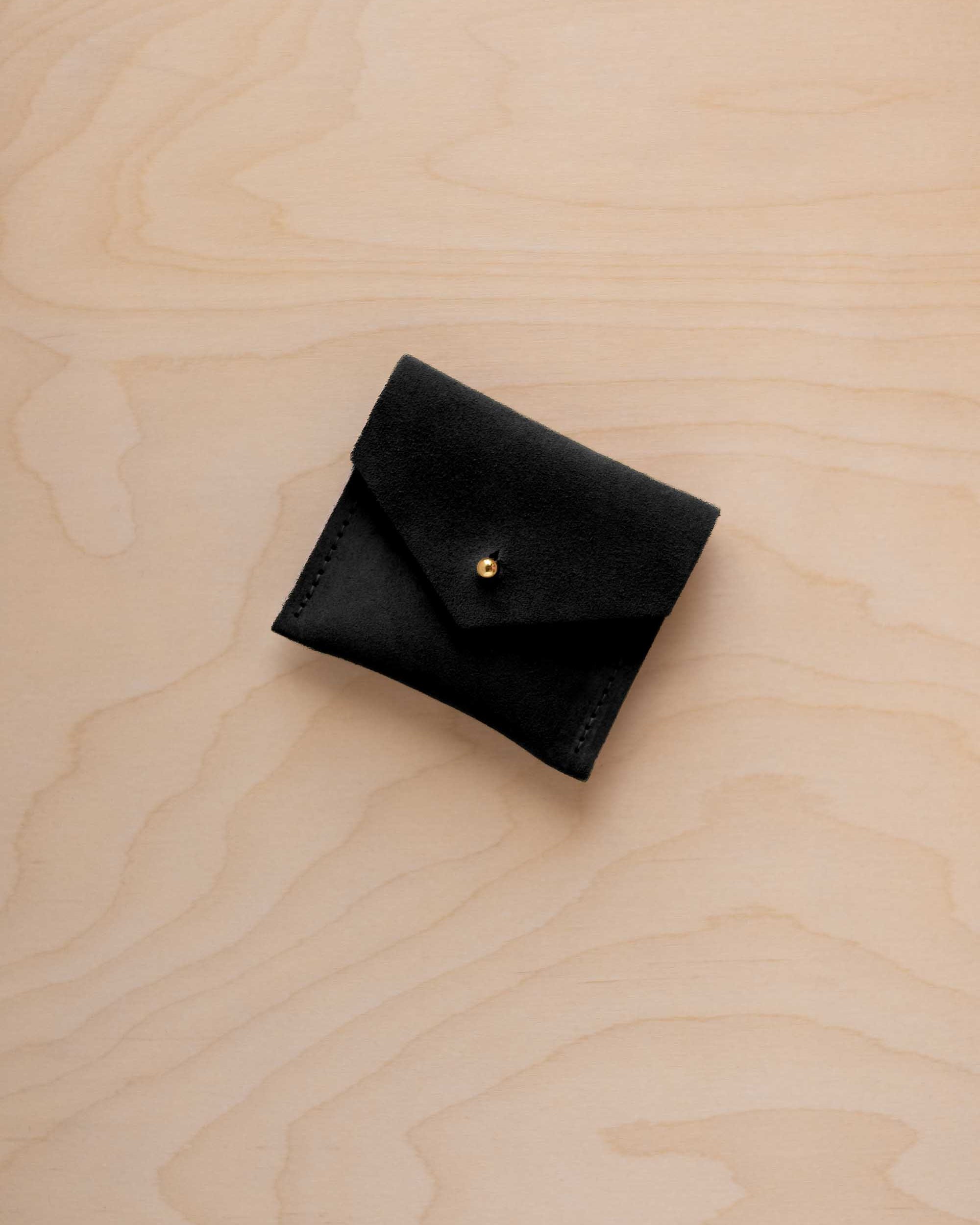 A small black suede pouch with a envelope flap closure and a gold button, placed on a light wood grain surface.