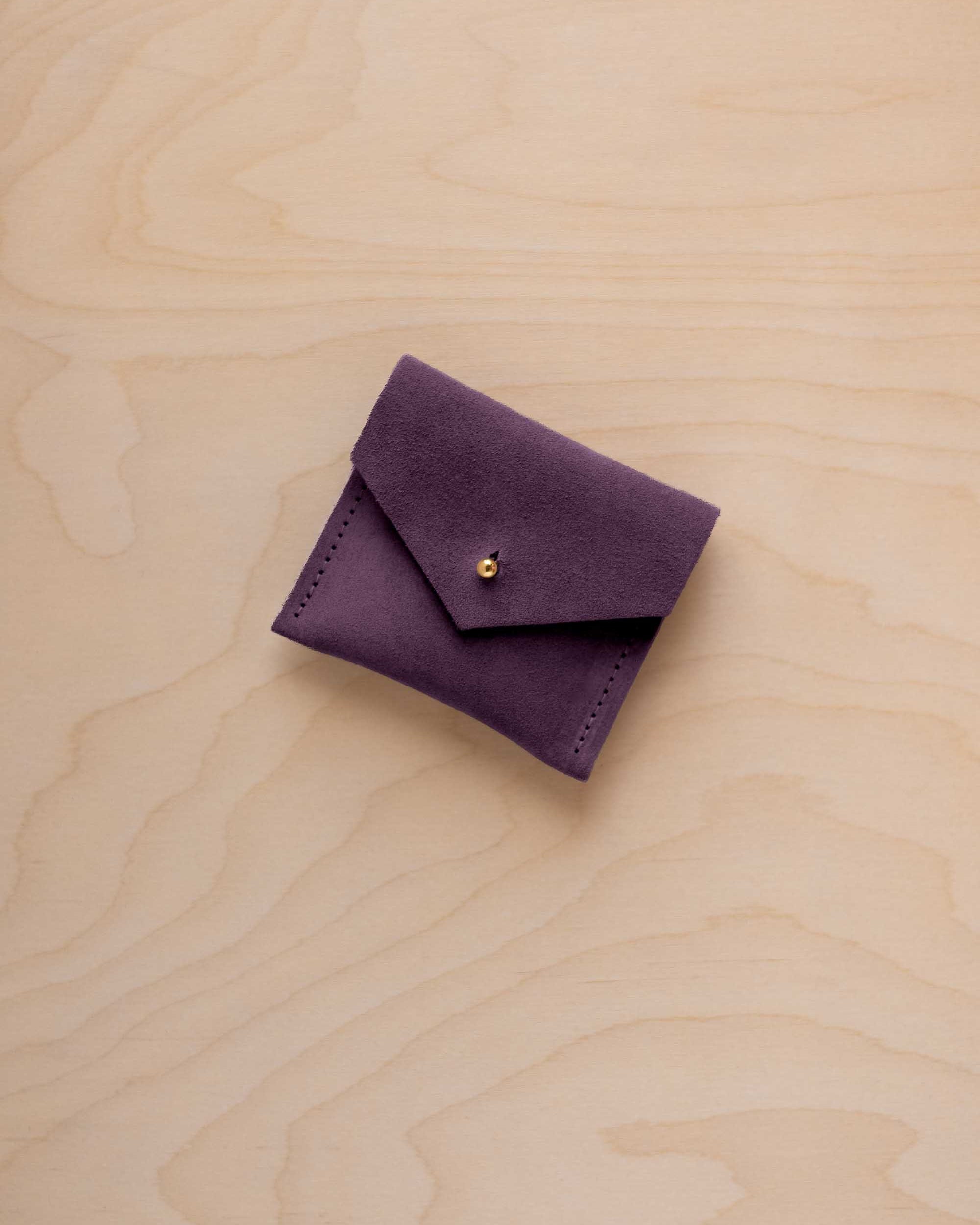 A small dark purple suede pouch with a flap closure and a gold button, placed on a light wood grain surface.