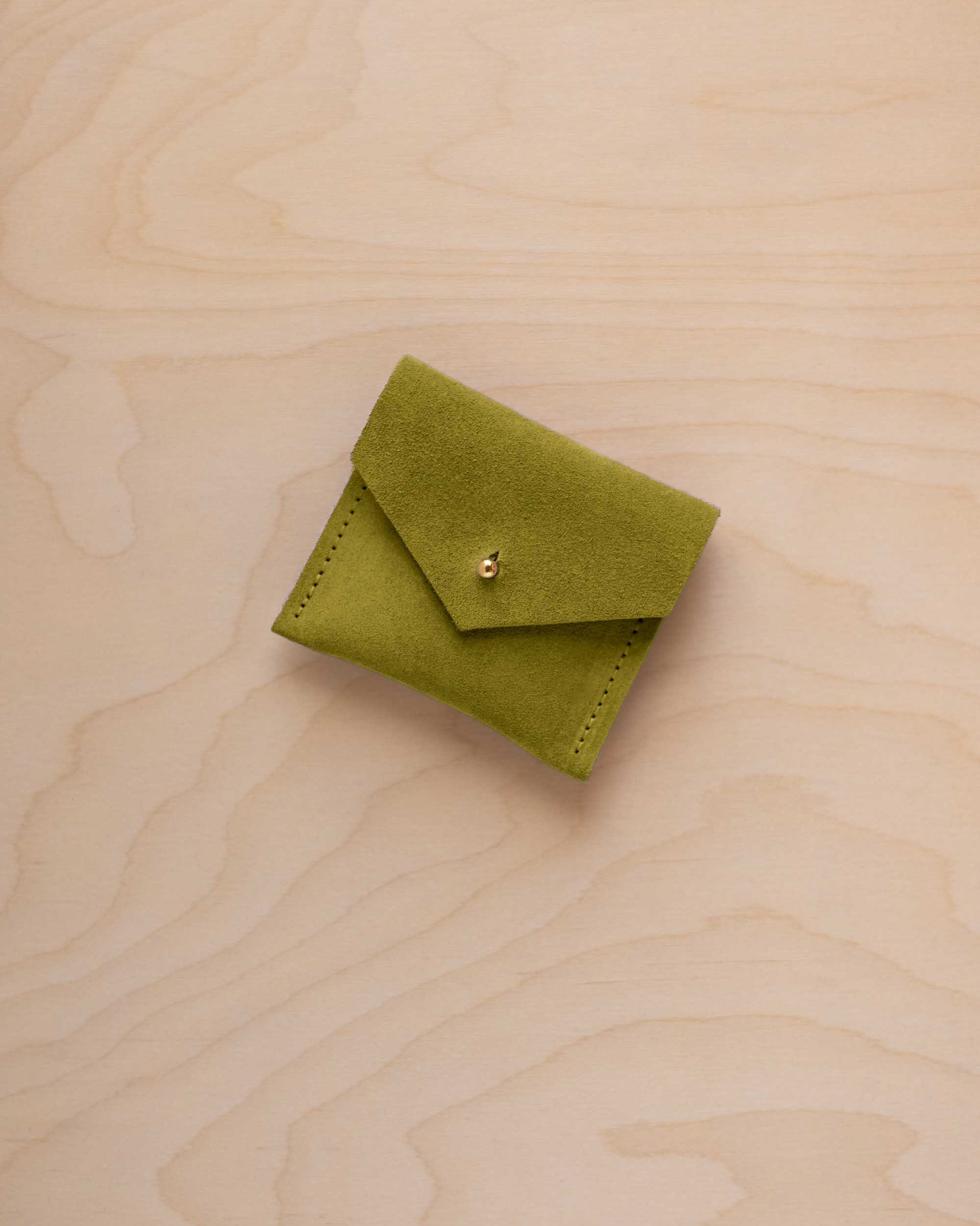 A small chartreuse green suede jewelry pouch with a gold button, resting on a light wood grain surface.