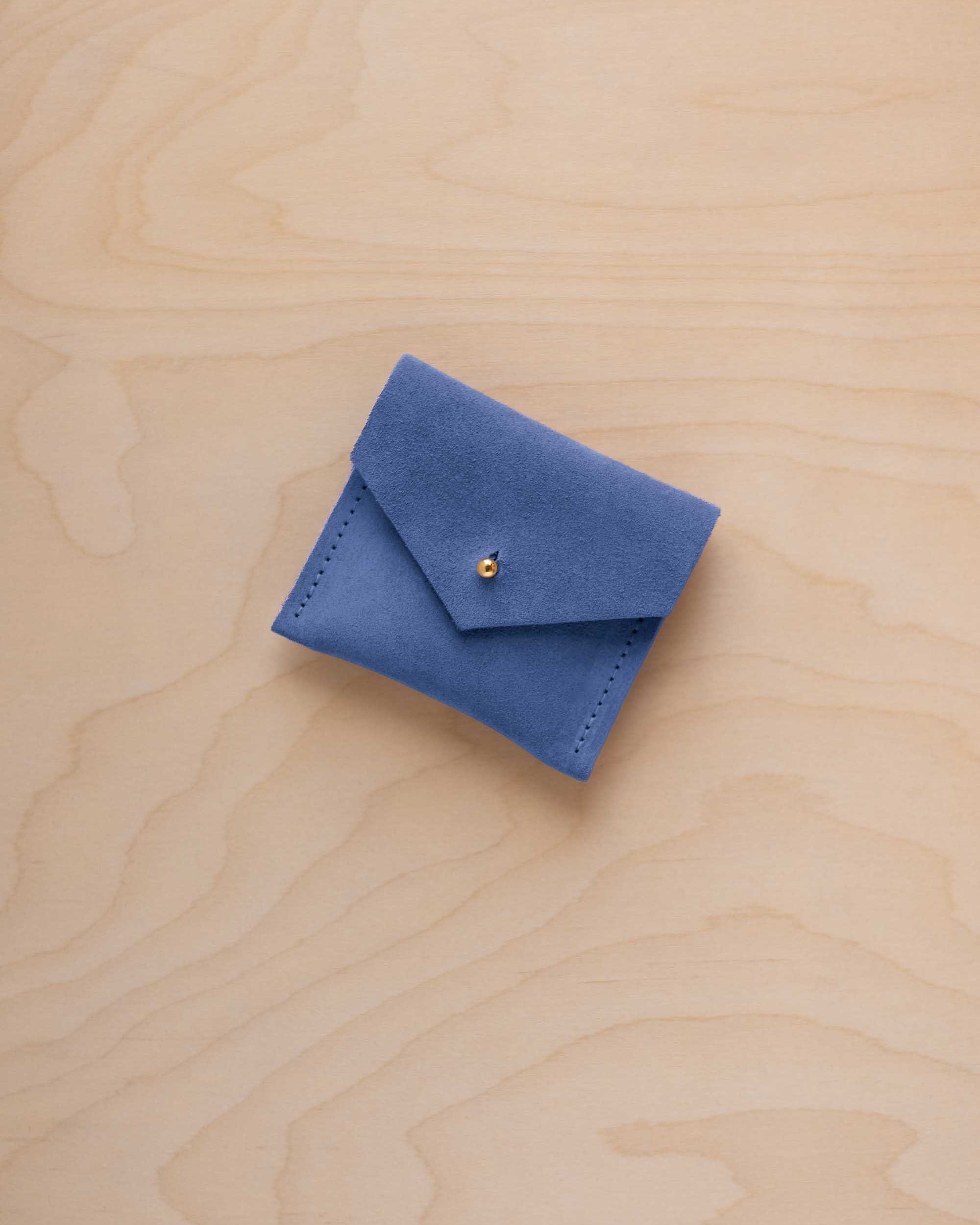 A small cobalt blue suede envelope-style pouch with a gold snap button, placed on a light wood grain surface.