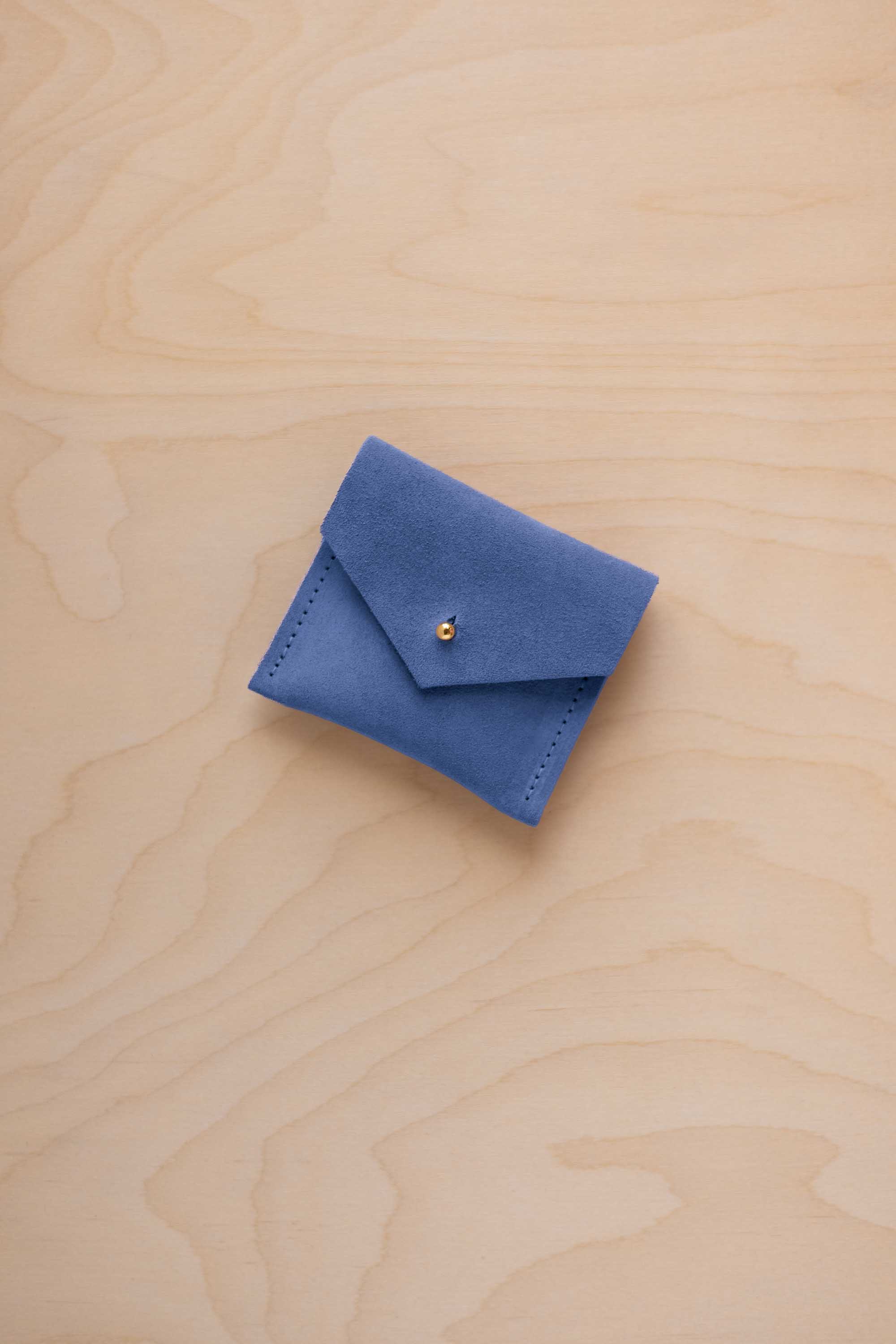A small cobalt blue suede envelope-style pouch with a gold snap button, placed on a light wood grain surface.