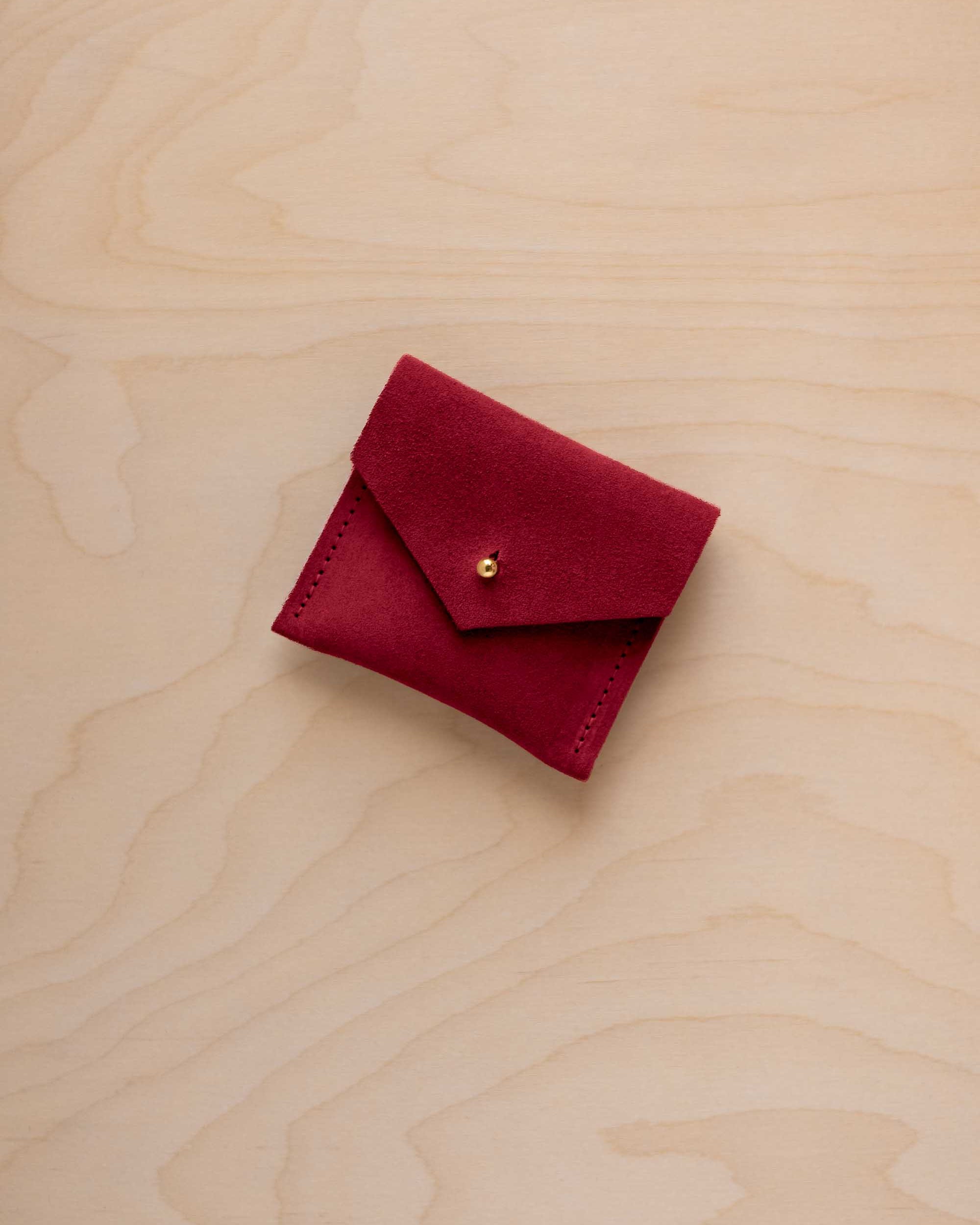 A small deep red suede pouch with a flap closure and a gold button, placed on a light wood grain surface.