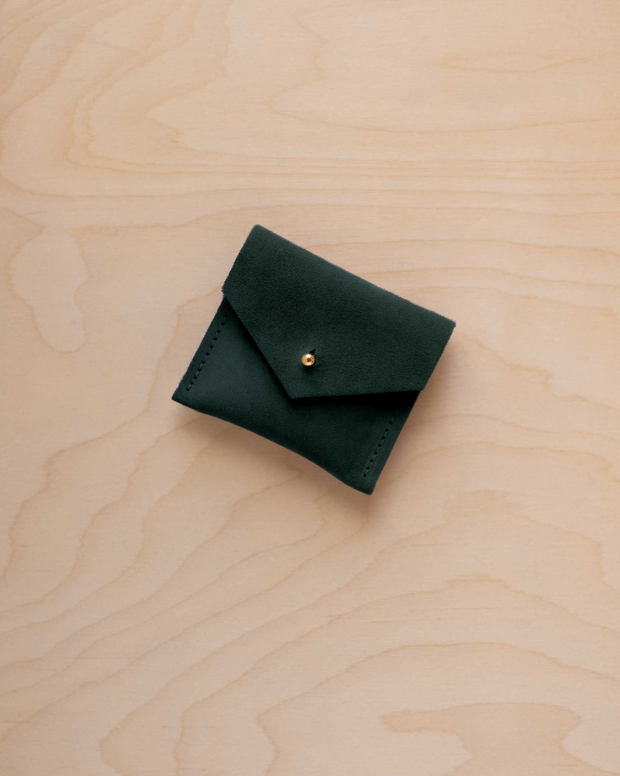 A small dark forest green suede pouch with a envelope flap closure and a gold button, placed on a light wood grain surface.