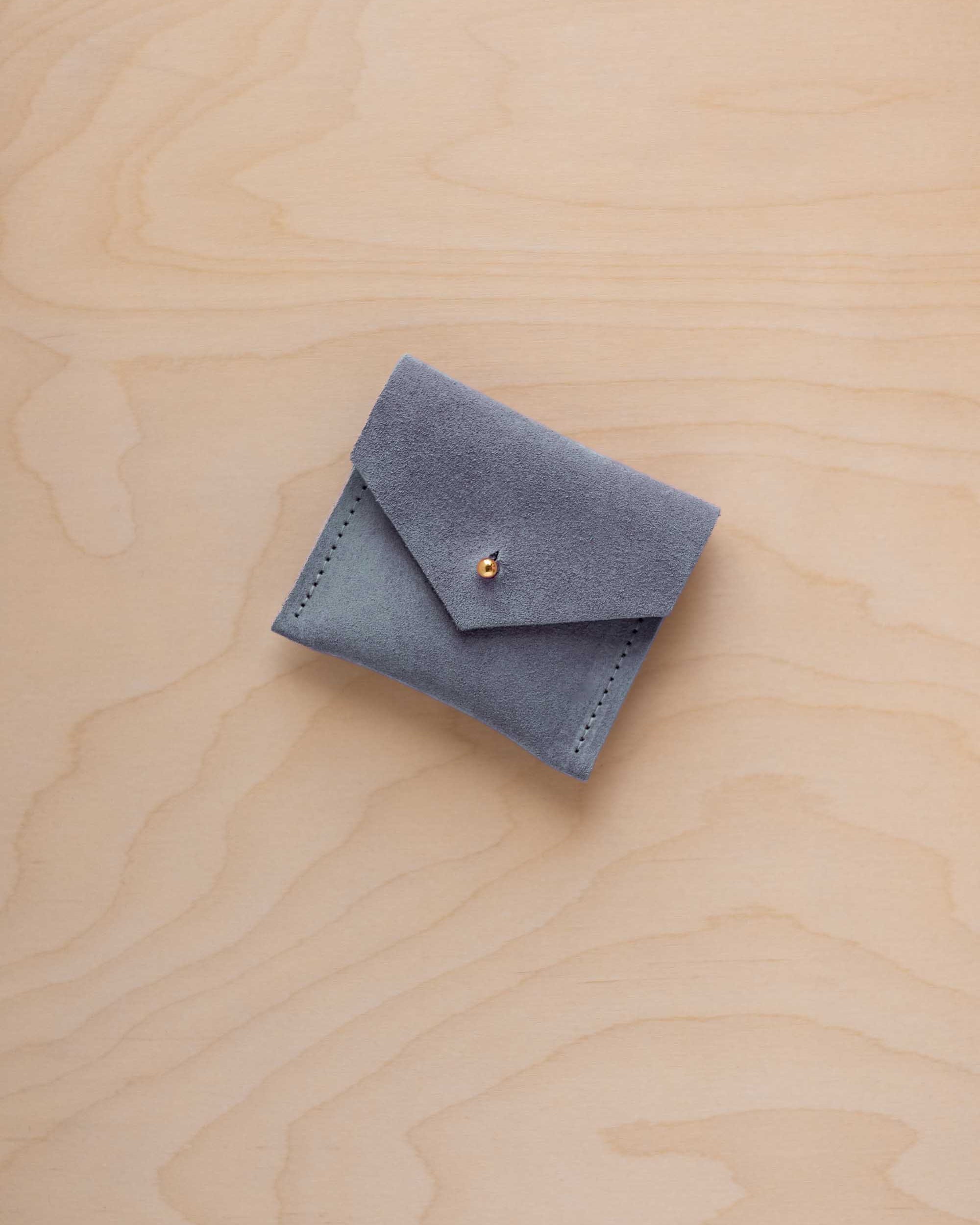 A small blue-gray suede jewelry pouch with a flap closure and a tiny gold button, placed on a light wood grain surface.
