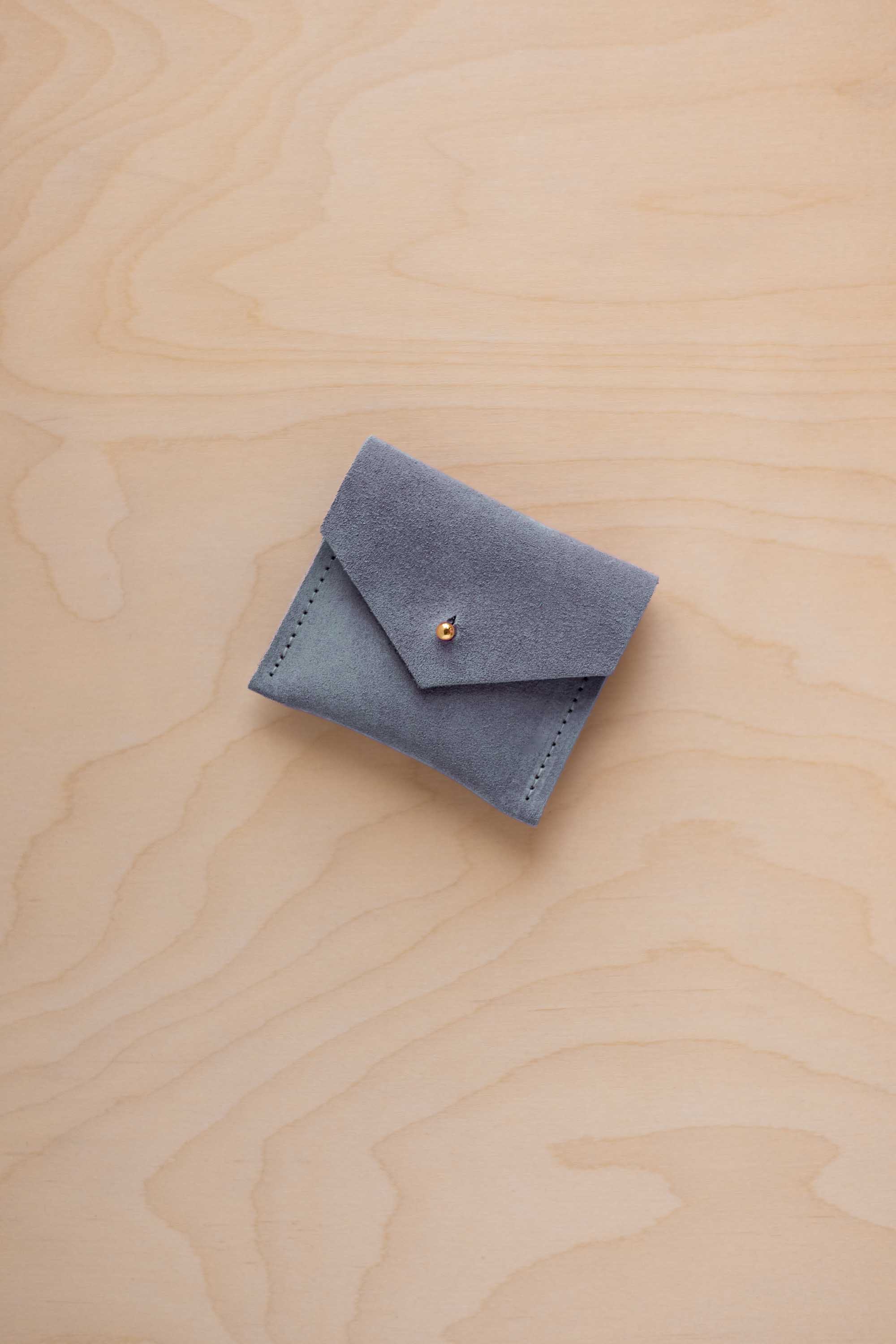 A small blue-gray suede jewelry pouch with a flap closure and a tiny gold button, placed on a light wood grain surface.