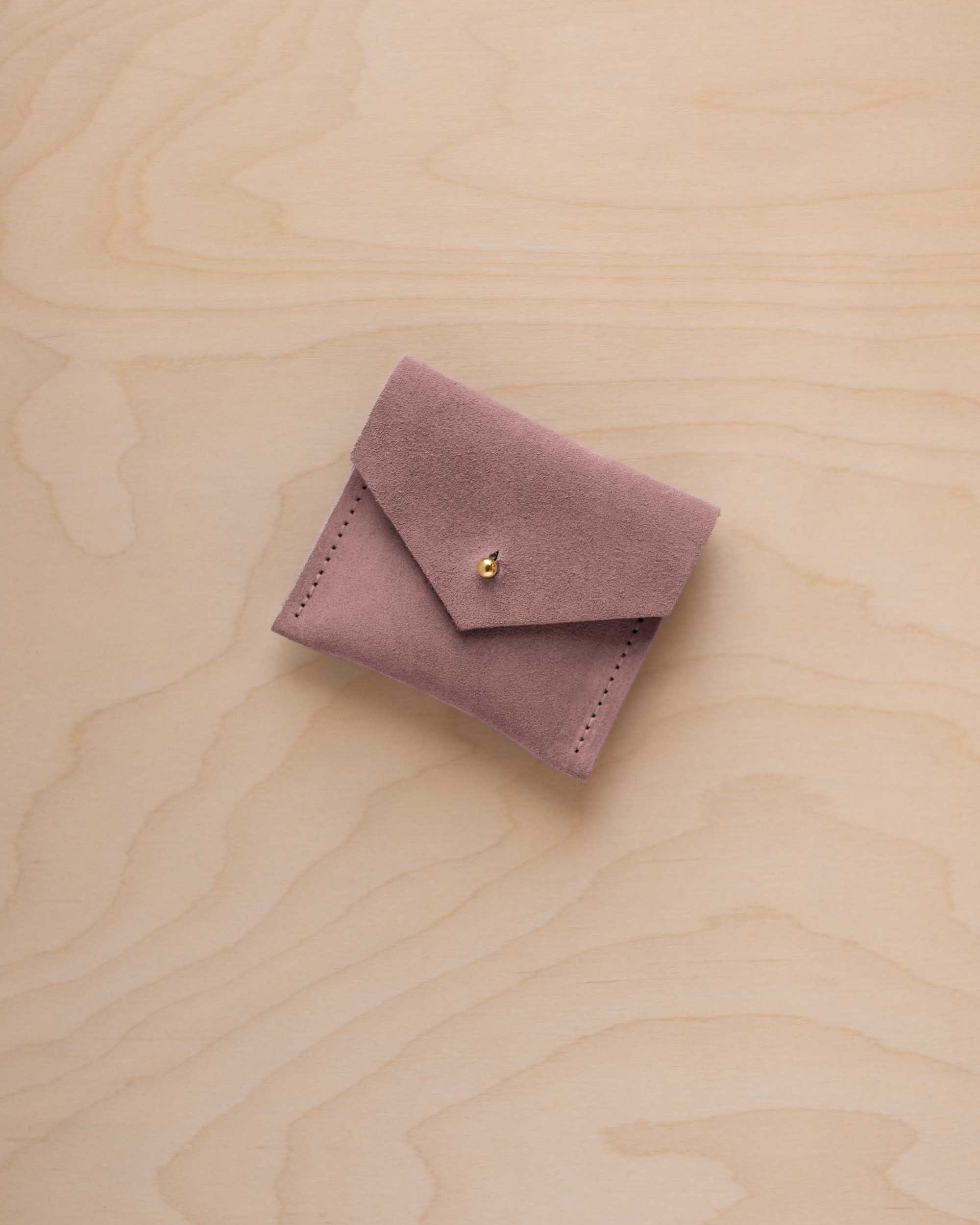 A small dusty mauve suede jewelry pouch with a flap closure and gold button, resting on a light wood grain background.