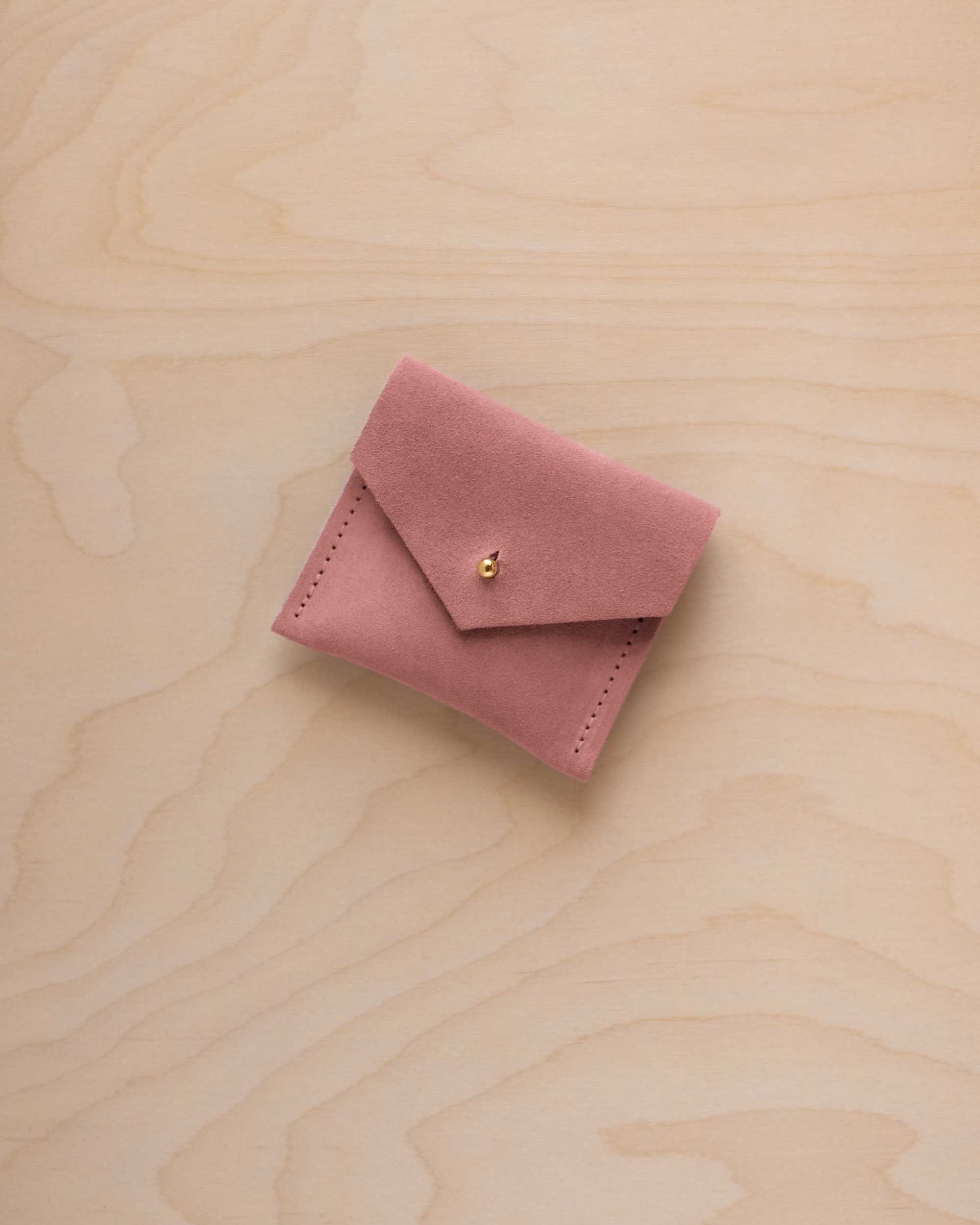 A small dusty pink suede envelope-style pouch with a gold button, resting on a light wood grain surface.