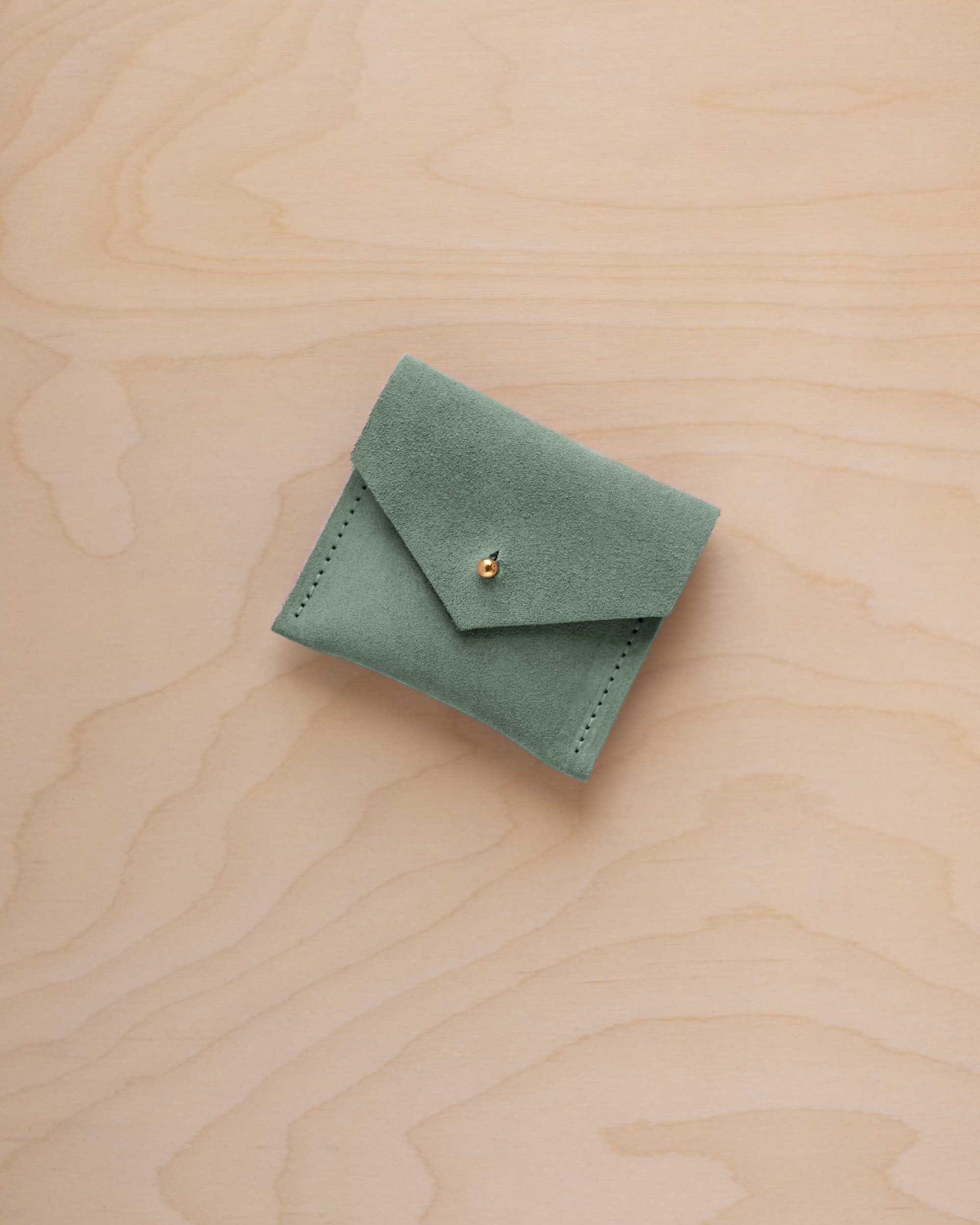 A small sage green suede jewelry pouch with a gold button fastening, resting on a light wood grain surface.