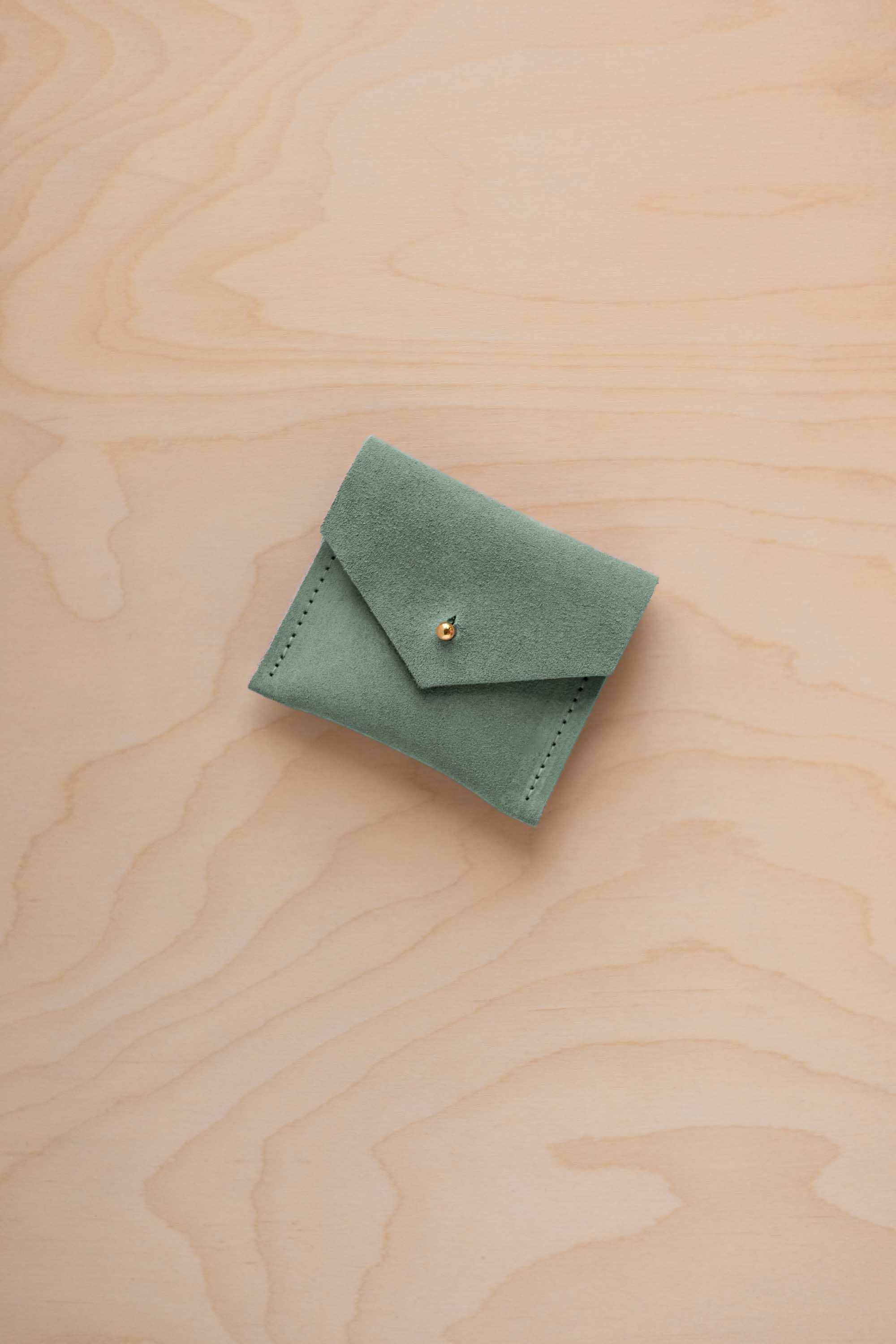A small sage green suede jewelry pouch with a gold button fastening, resting on a light wood grain surface.