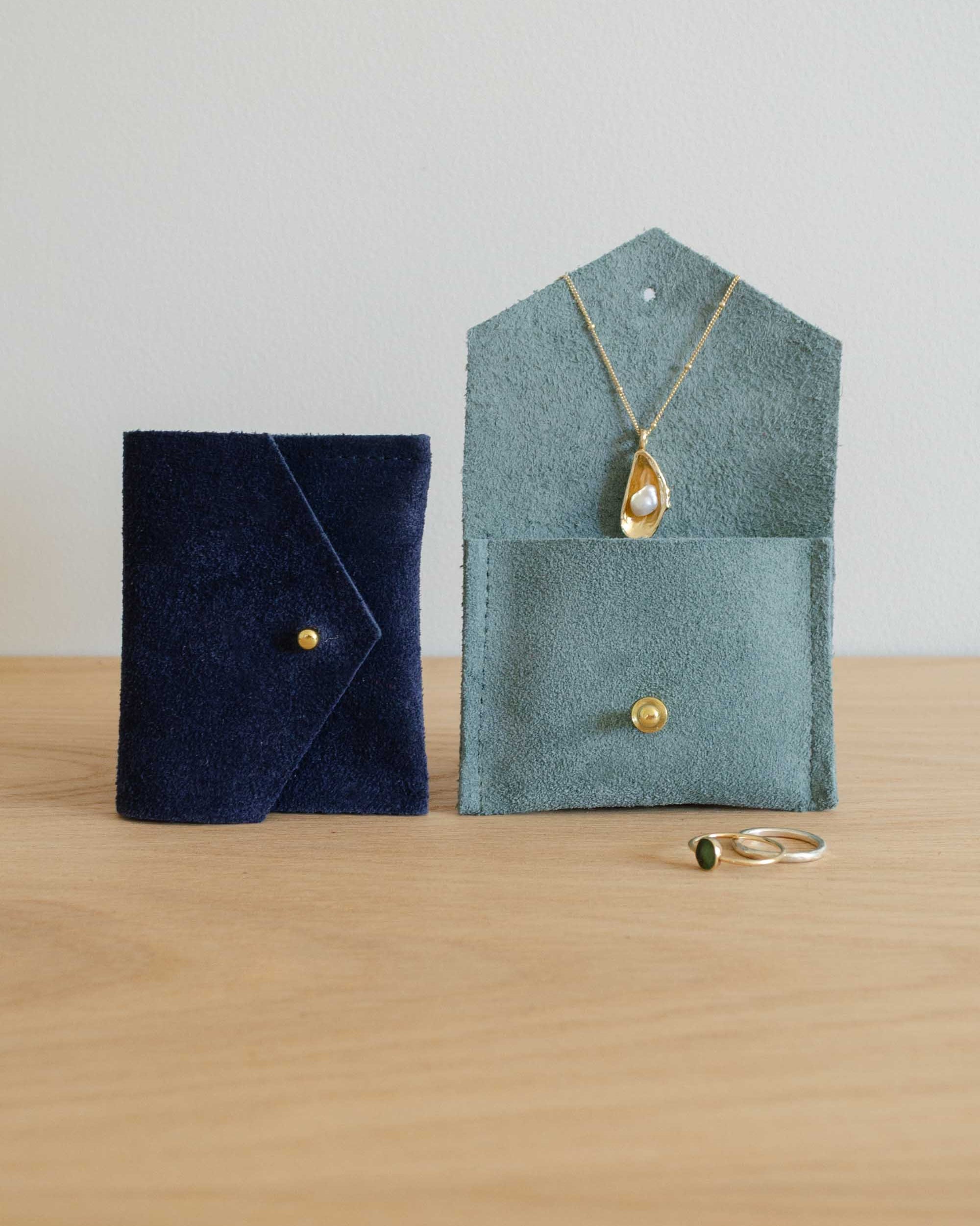 Two small suede jewelry pouches on a light wood surface against a plain light gray wall. One pouch is dark navy blue and the other is muted sage green, both with a button closure.
