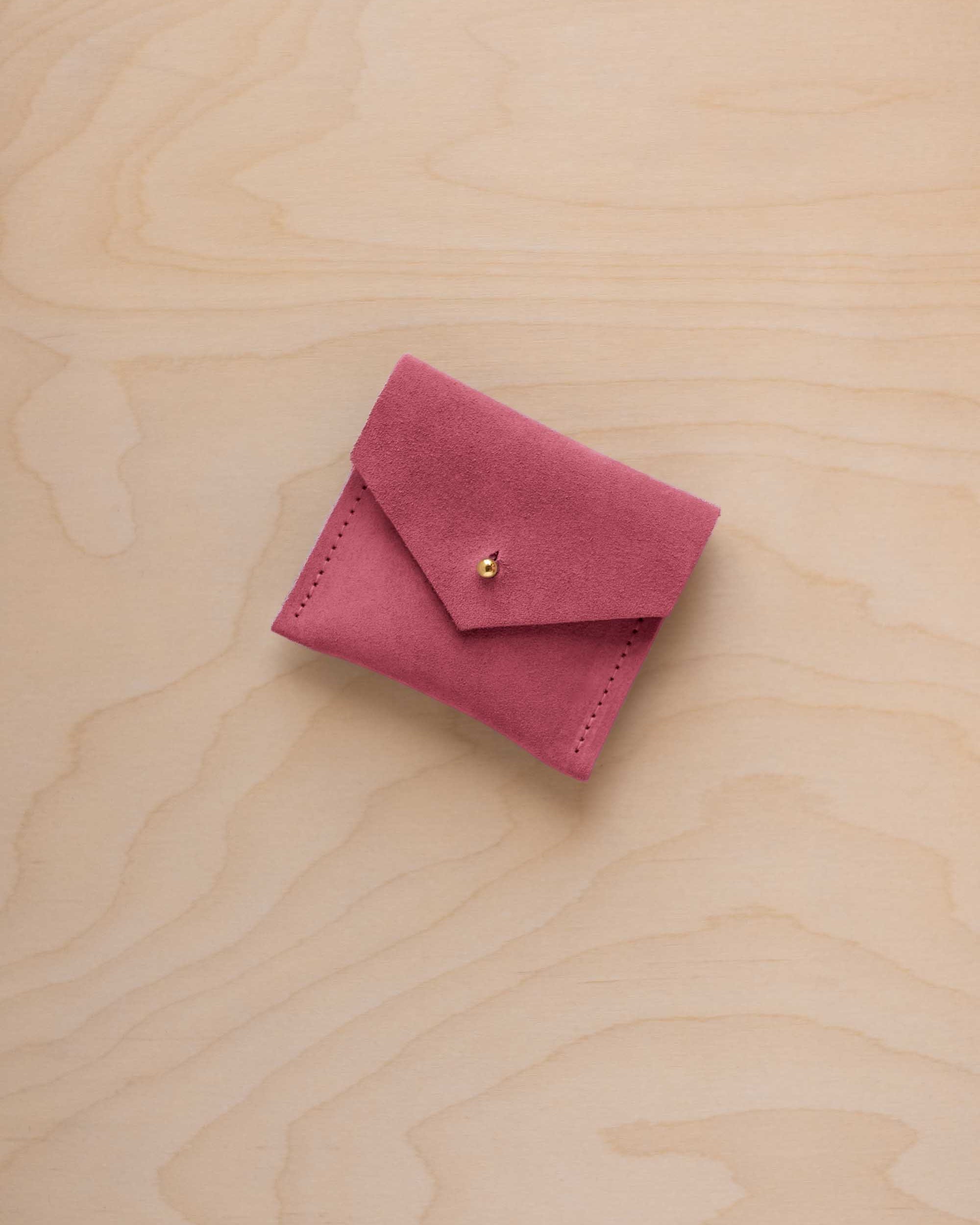 A small pink suede pouch with a flap closure and a tiny gold button, placed on a light wood grain surface.