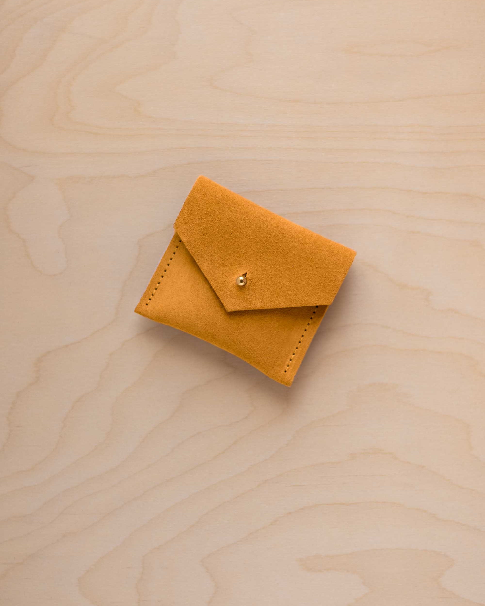 A small mustard yellow suede envelope-style pouch with a tiny gold button, placed on a light wood grain surface.