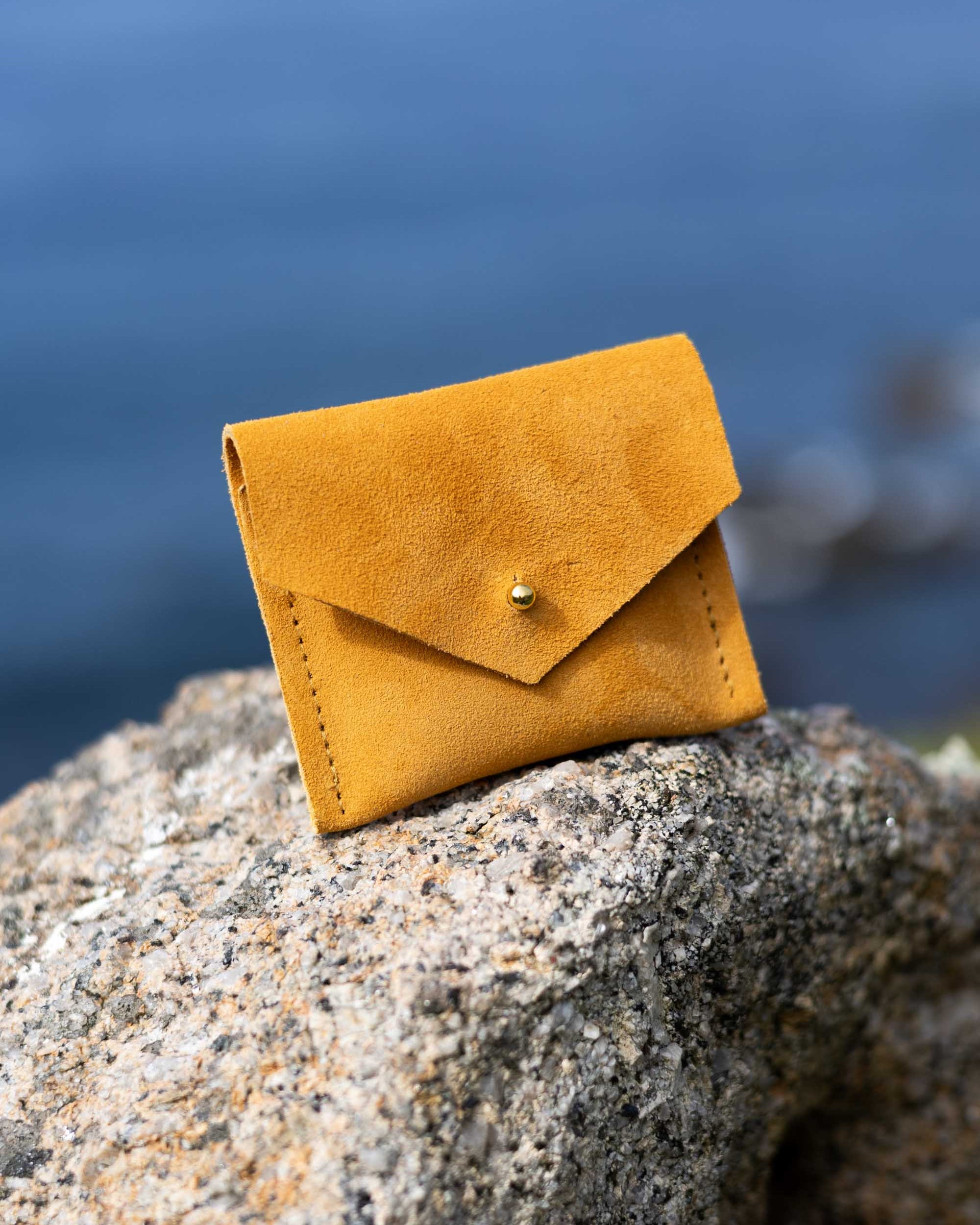 A small mustard yellow suede envelope-style card holder or wallet with a gold button, resting on a textured gray rock with a blurred blue water background.
