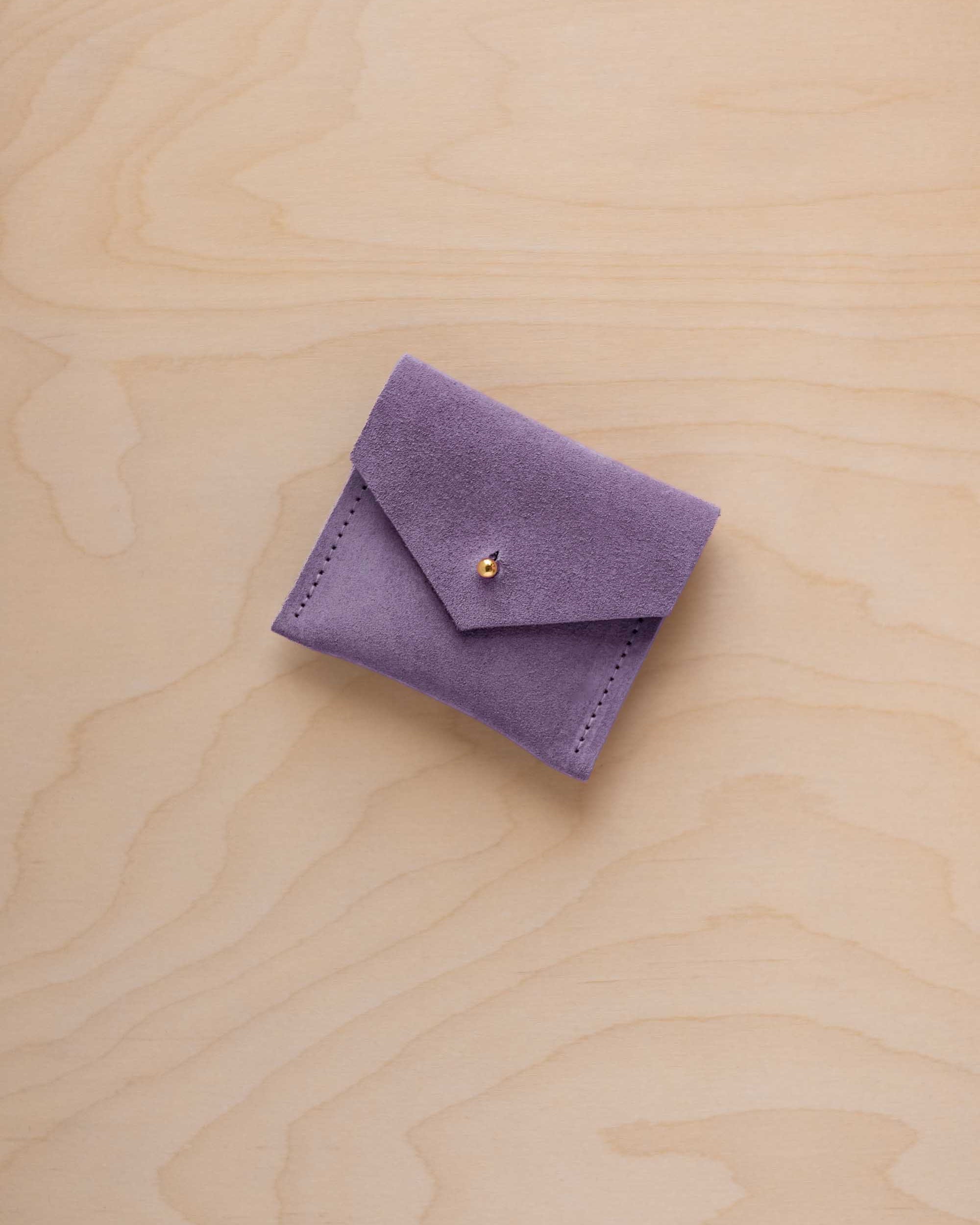 A small lavender purple suede jewelry pouch with a gold button, resting on a light wood grain background.