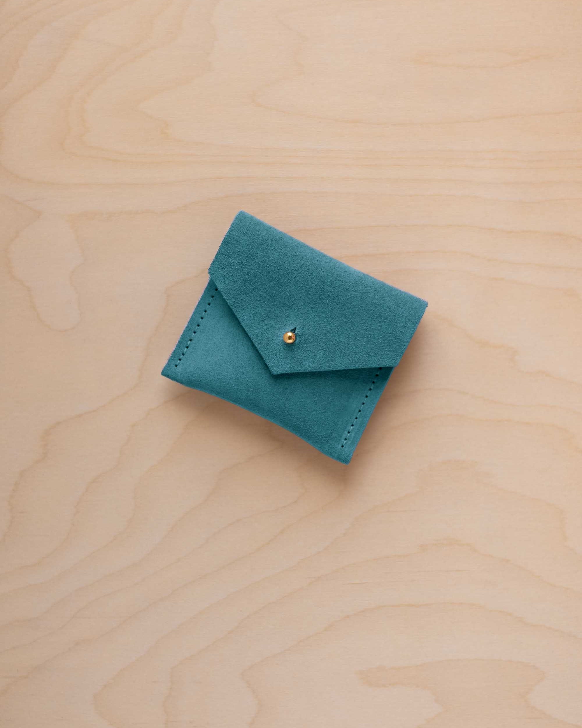 A small teal blue suede jewelry pouch with a gold button, resting on a light wood grain surface.