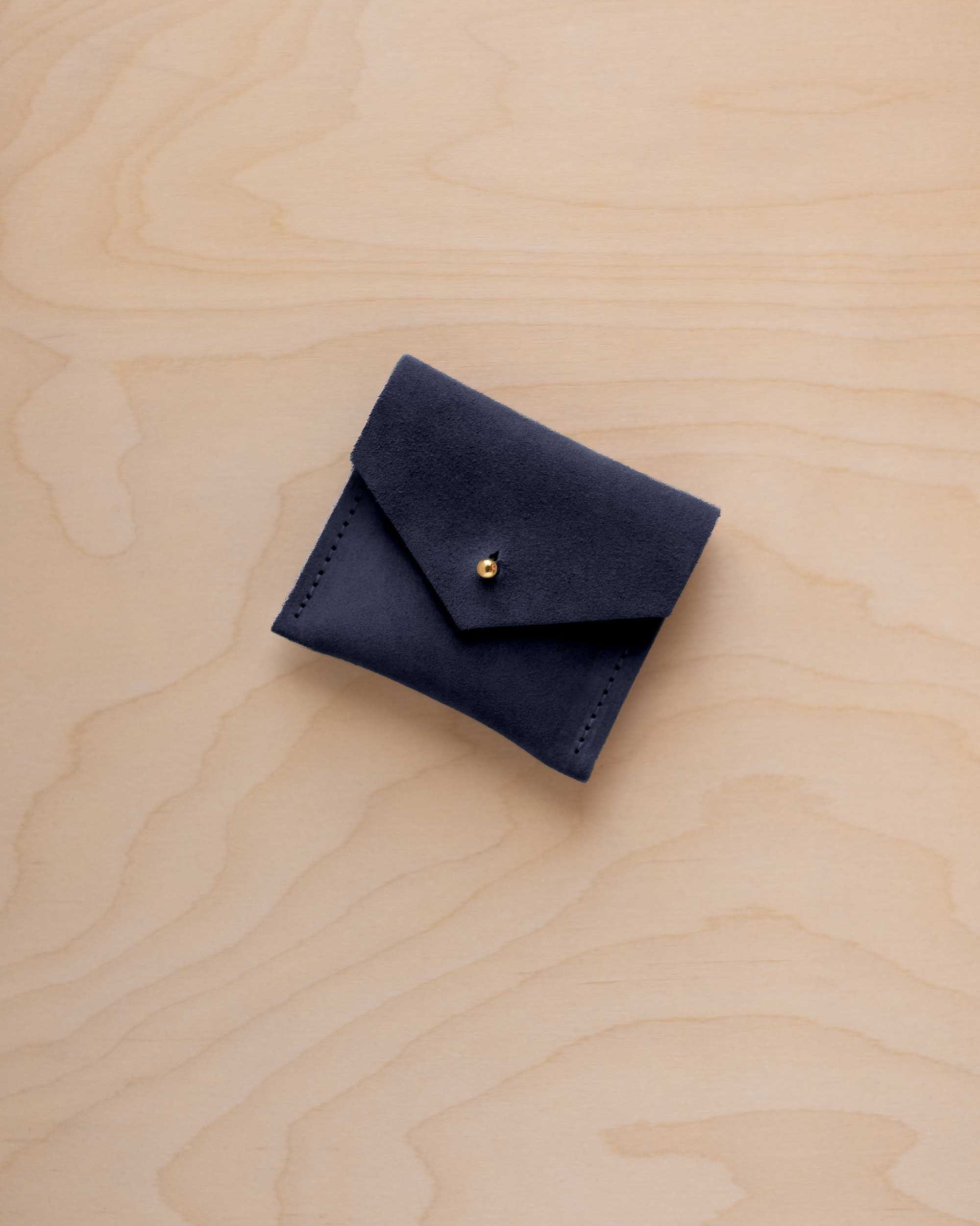A small dark navy suede pouch with a flap closure and a gold-tone button, placed on a light wood grain surface.