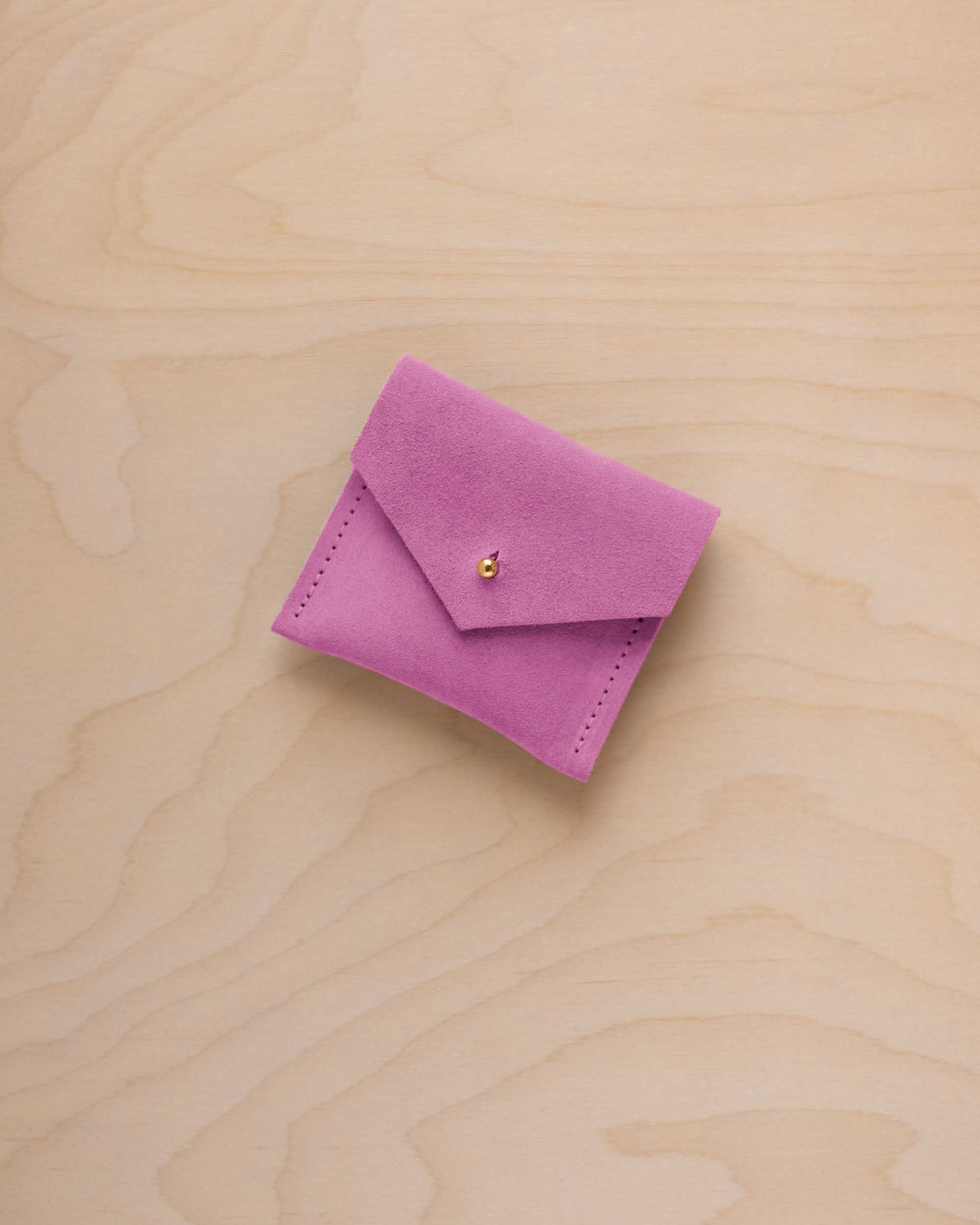 A small pink suede pouch with a flap closure and a tiny gold coloured button, placed on a light wood grain surface.