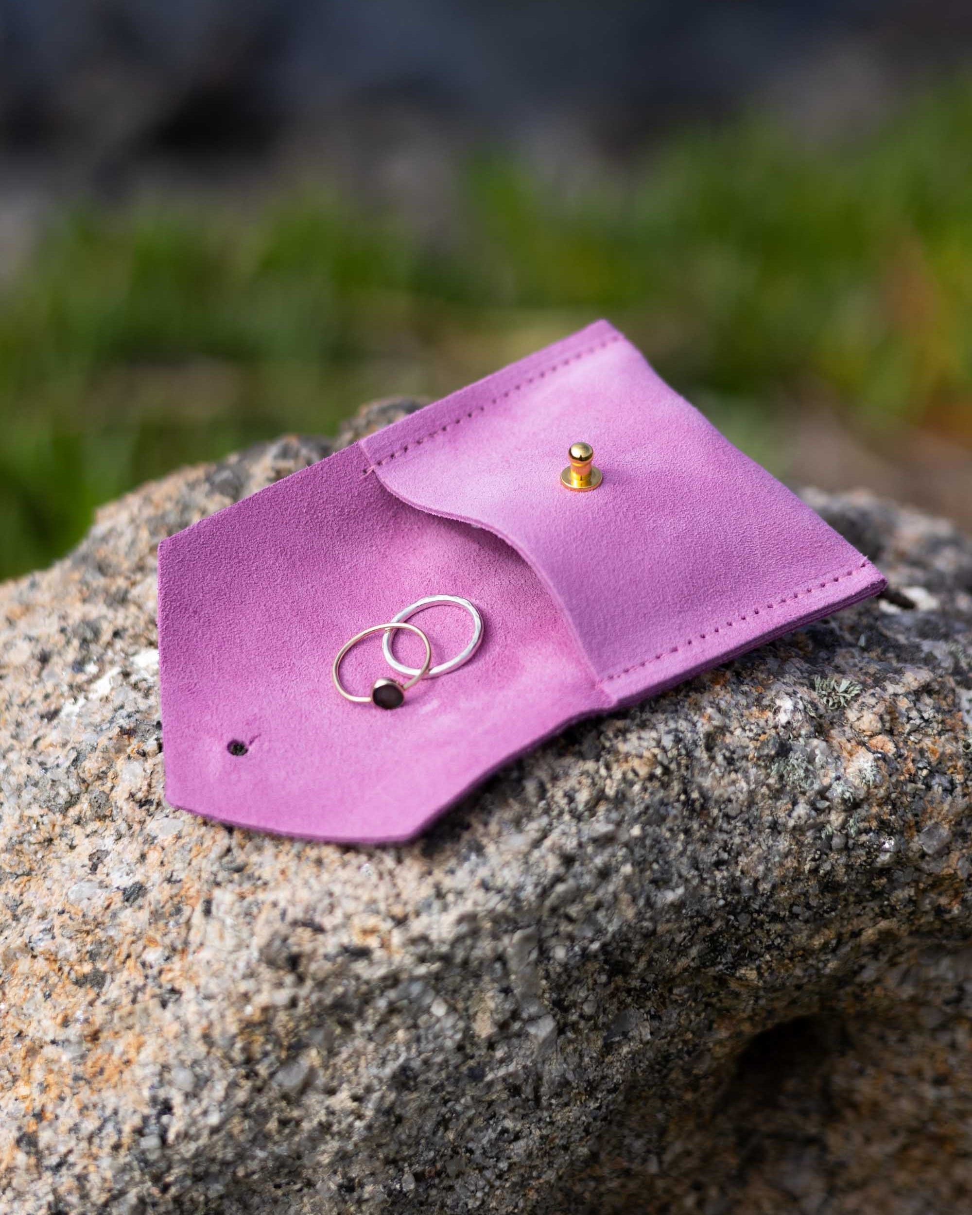 A small pink leather jewelry pouch or ring holder resting on a rough stone outdoors, with two rings placed inside and a small gold stud earring on the flap.