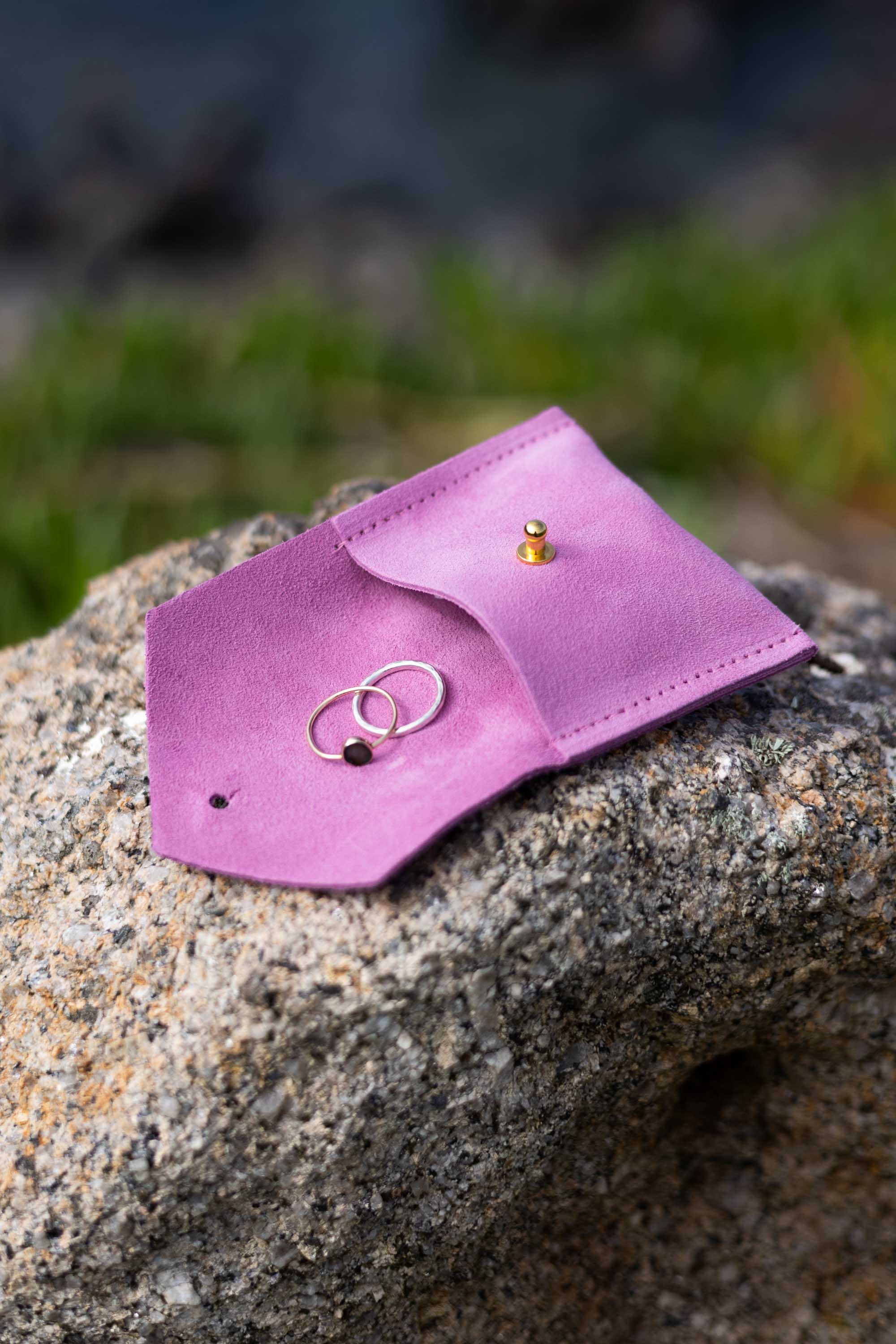 A small pink leather jewelry pouch or ring holder resting on a rough stone outdoors, with two rings placed inside and a small gold stud earring on the flap.