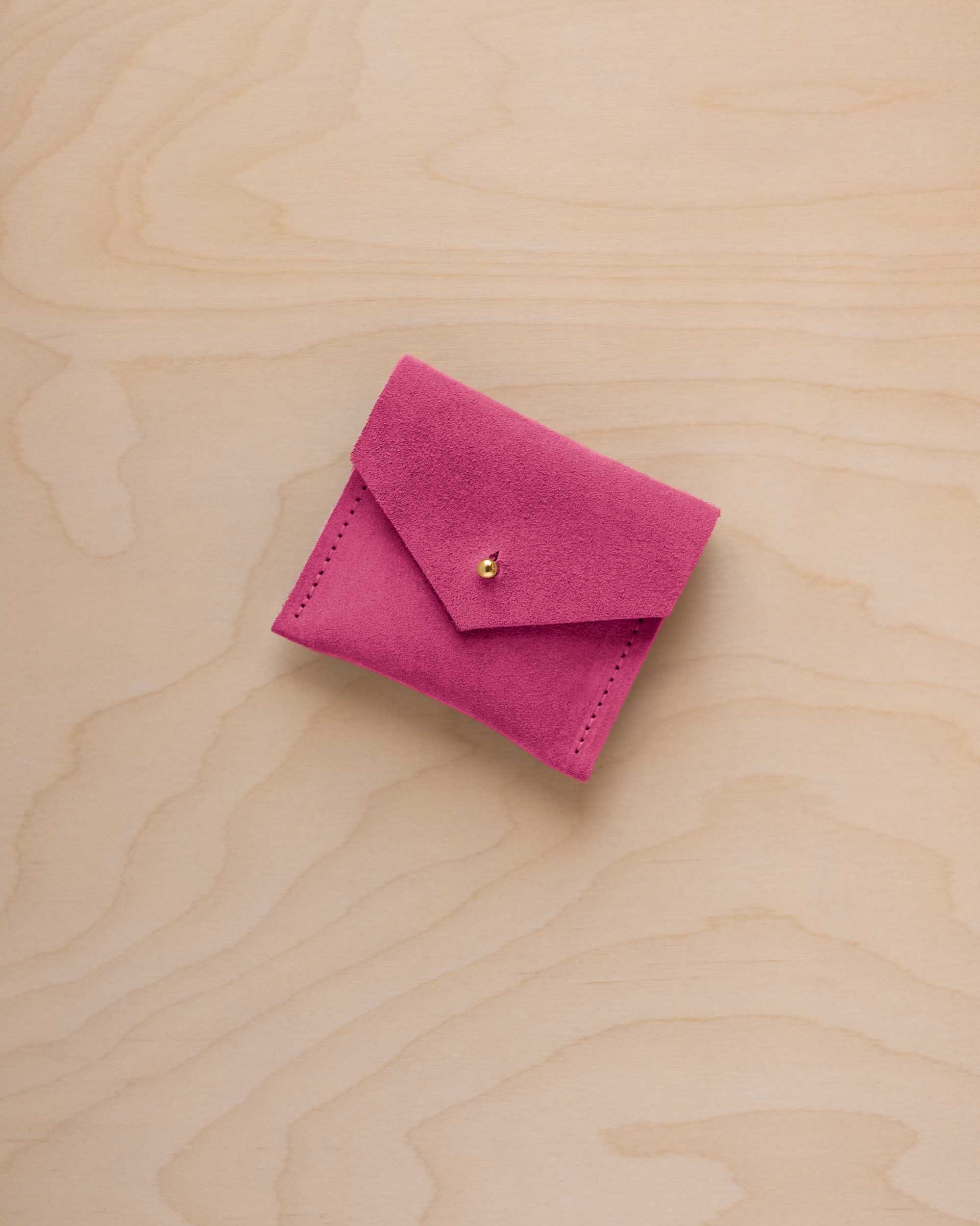 A small bright pink suede pouch with a flap closure and a tiny gold button, placed on a light wood grain surface.