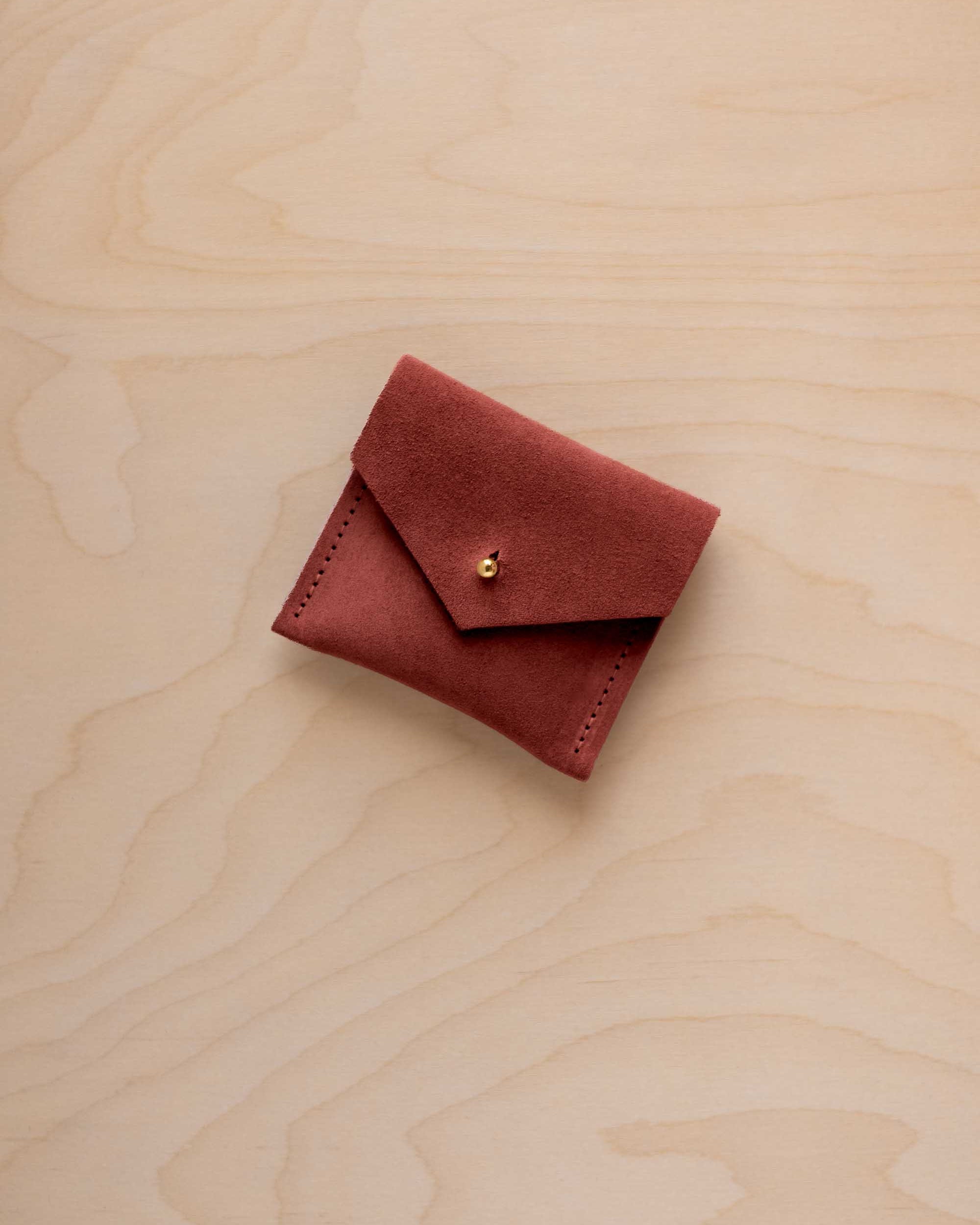 A small reddish-brown suede pouch with a flap closure and a gold button, placed on a light wood grain surface.