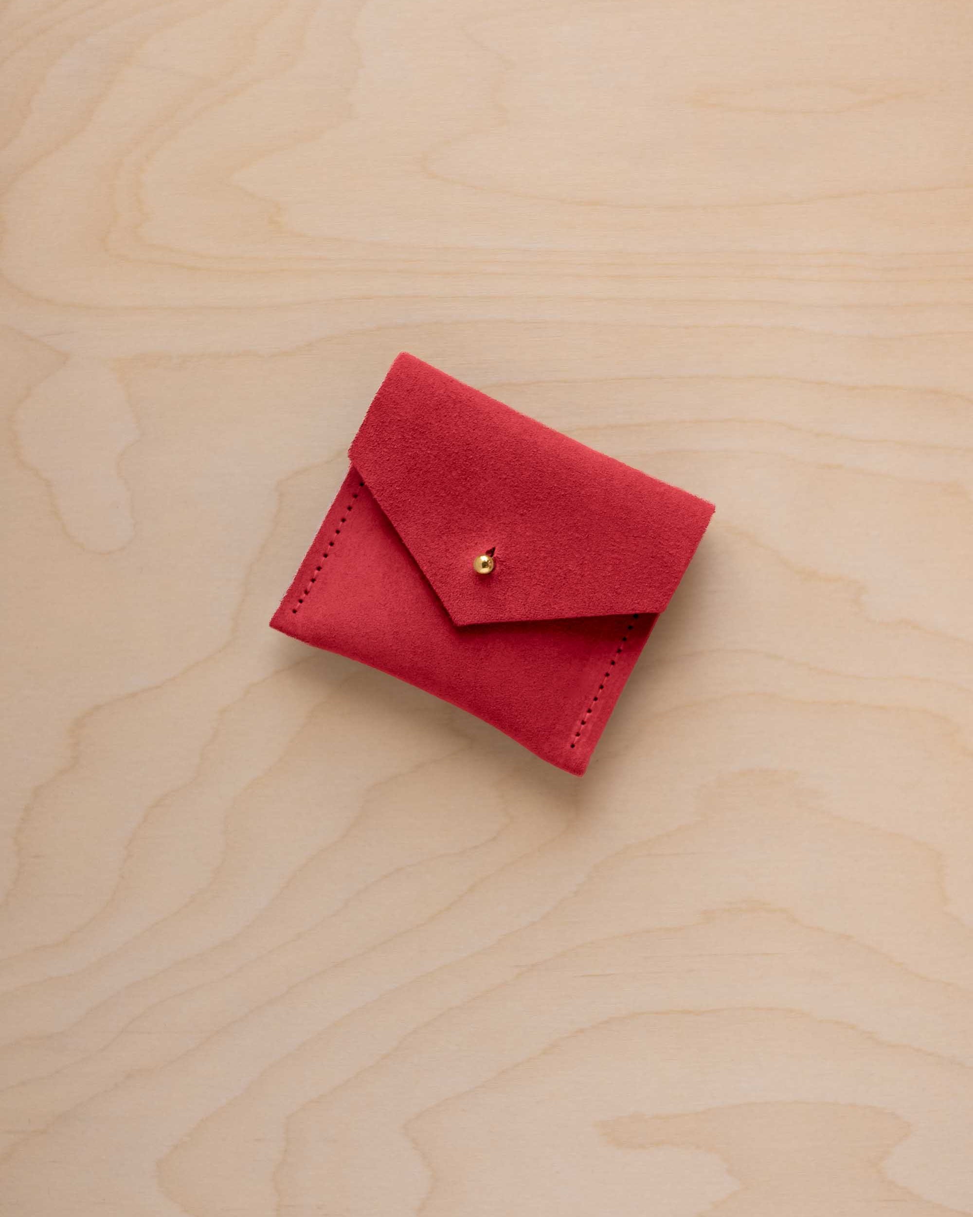 A small red suede pouch with a flap closure and a tiny gold button, placed on a light wood grain surface.