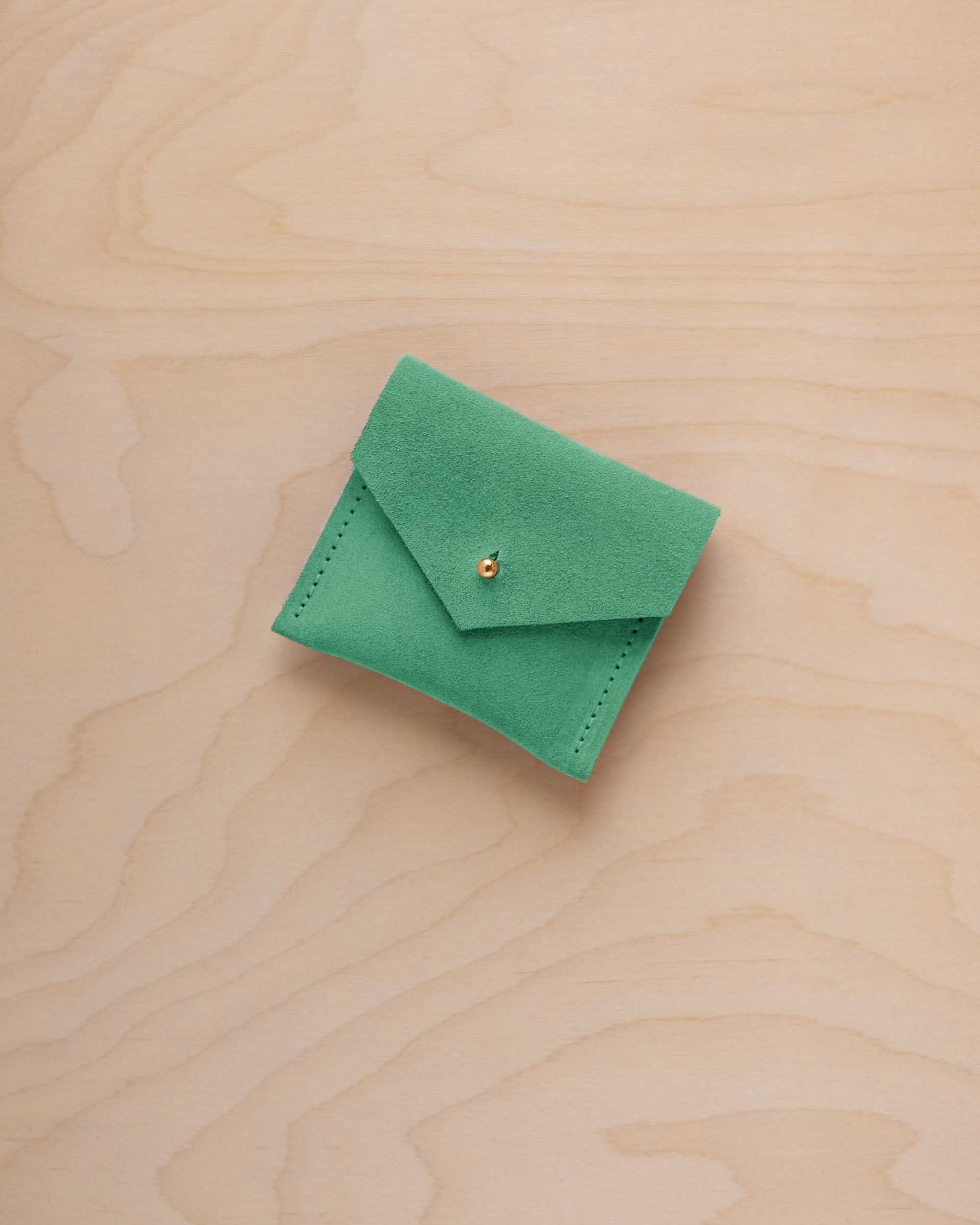 A small light emerald green suede pouch with a gold coloured button closure, placed on a light wood grain surface.