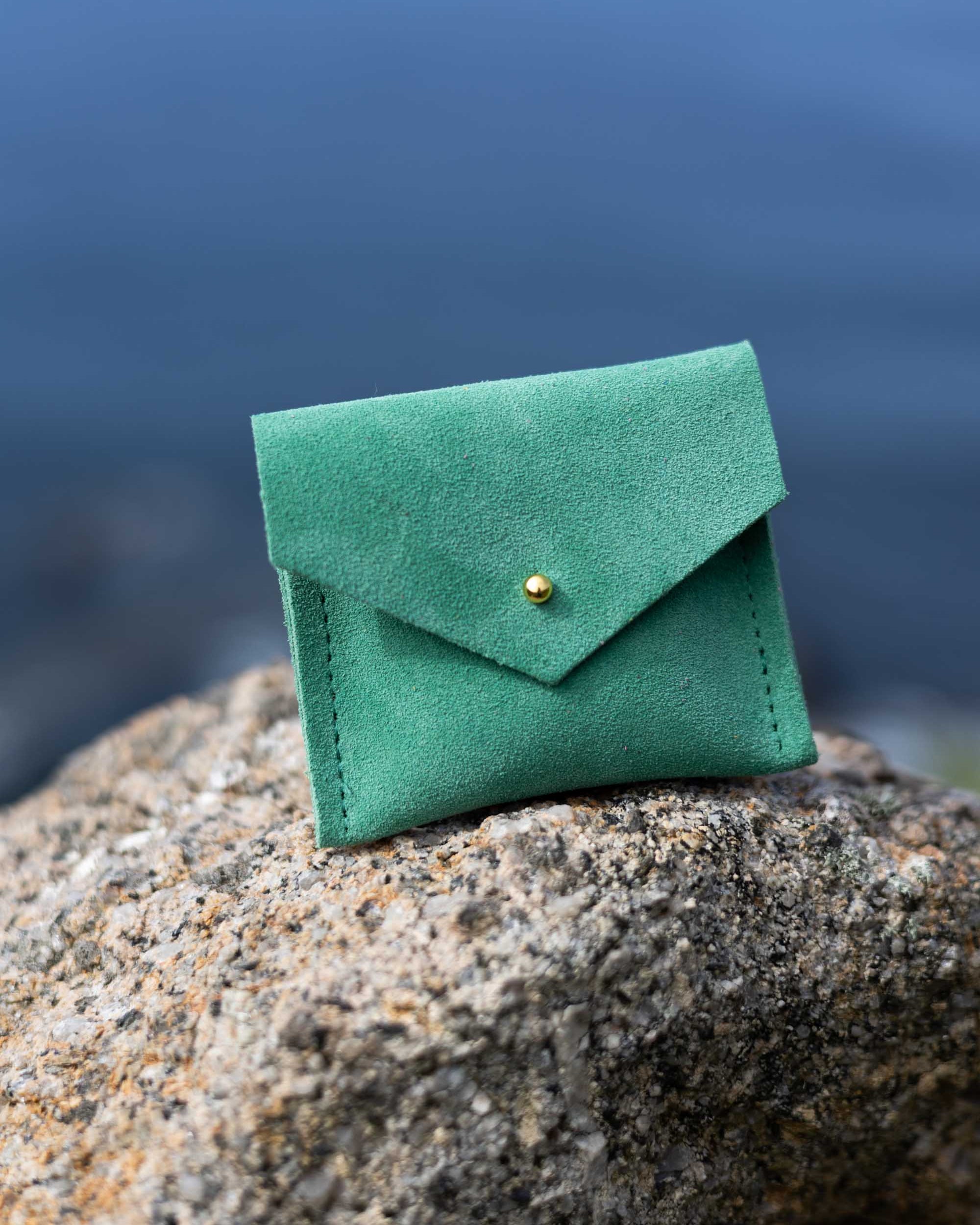 A small green suede envelope-style pouch with a gold button, resting on a large textured rock with a blurred outdoor background.