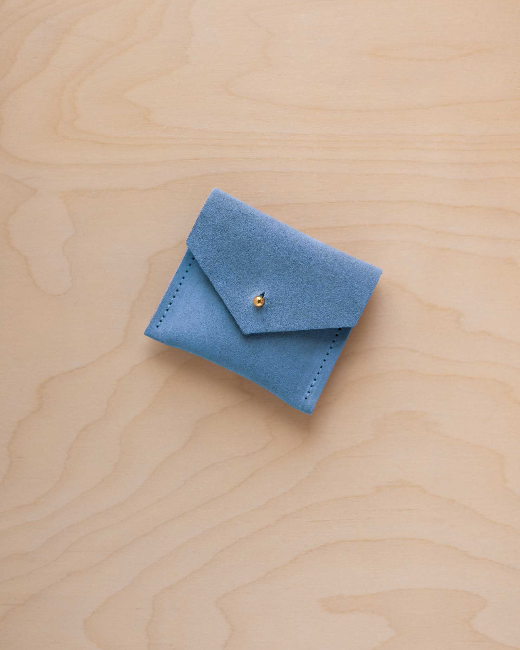 A small bright sky blue leather pouch with a flap closure and a tiny gold button, placed on a light wood grain surface.