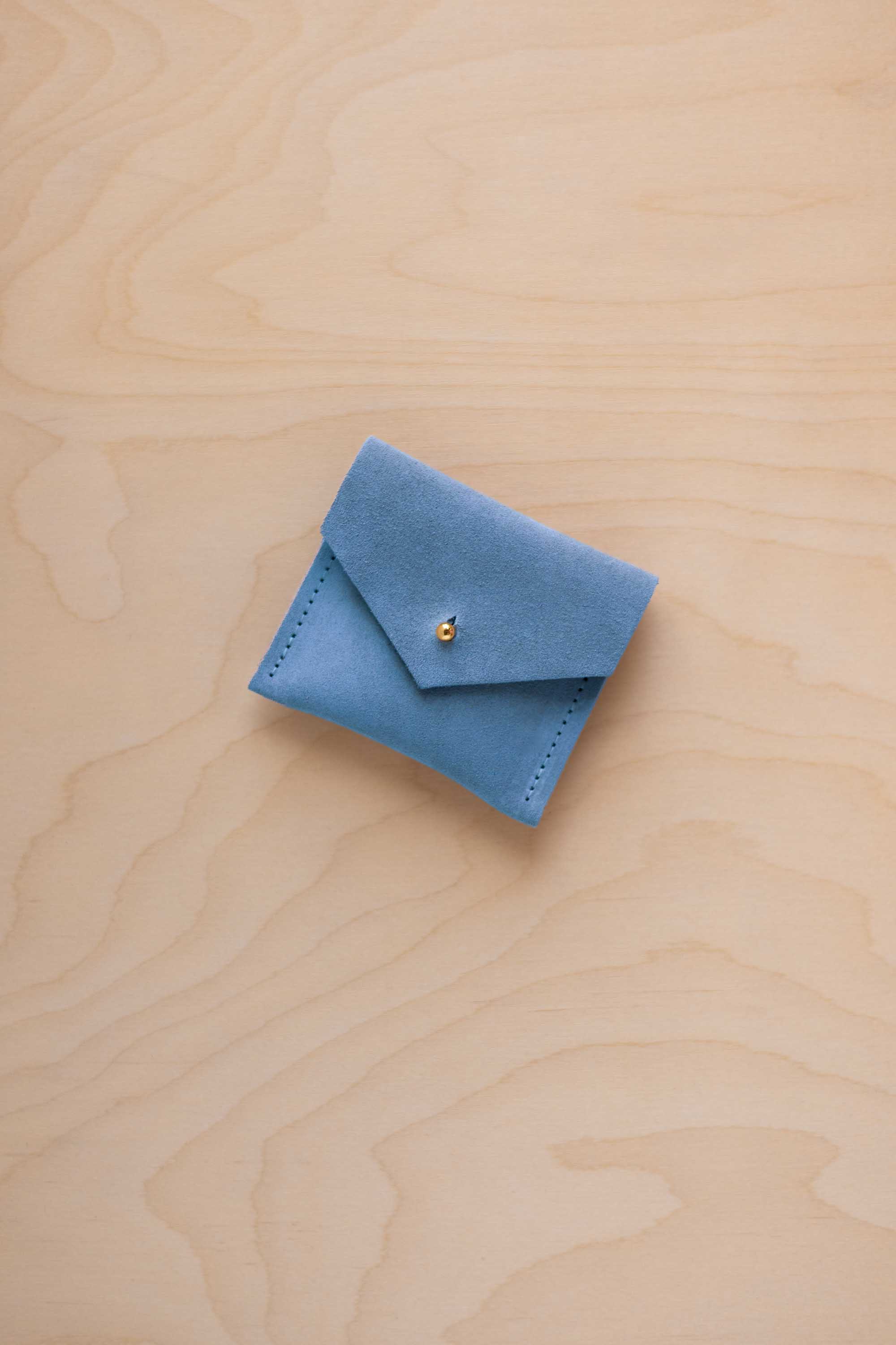 A small bright sky blue leather pouch with a flap closure and a tiny gold button, placed on a light wood grain surface.