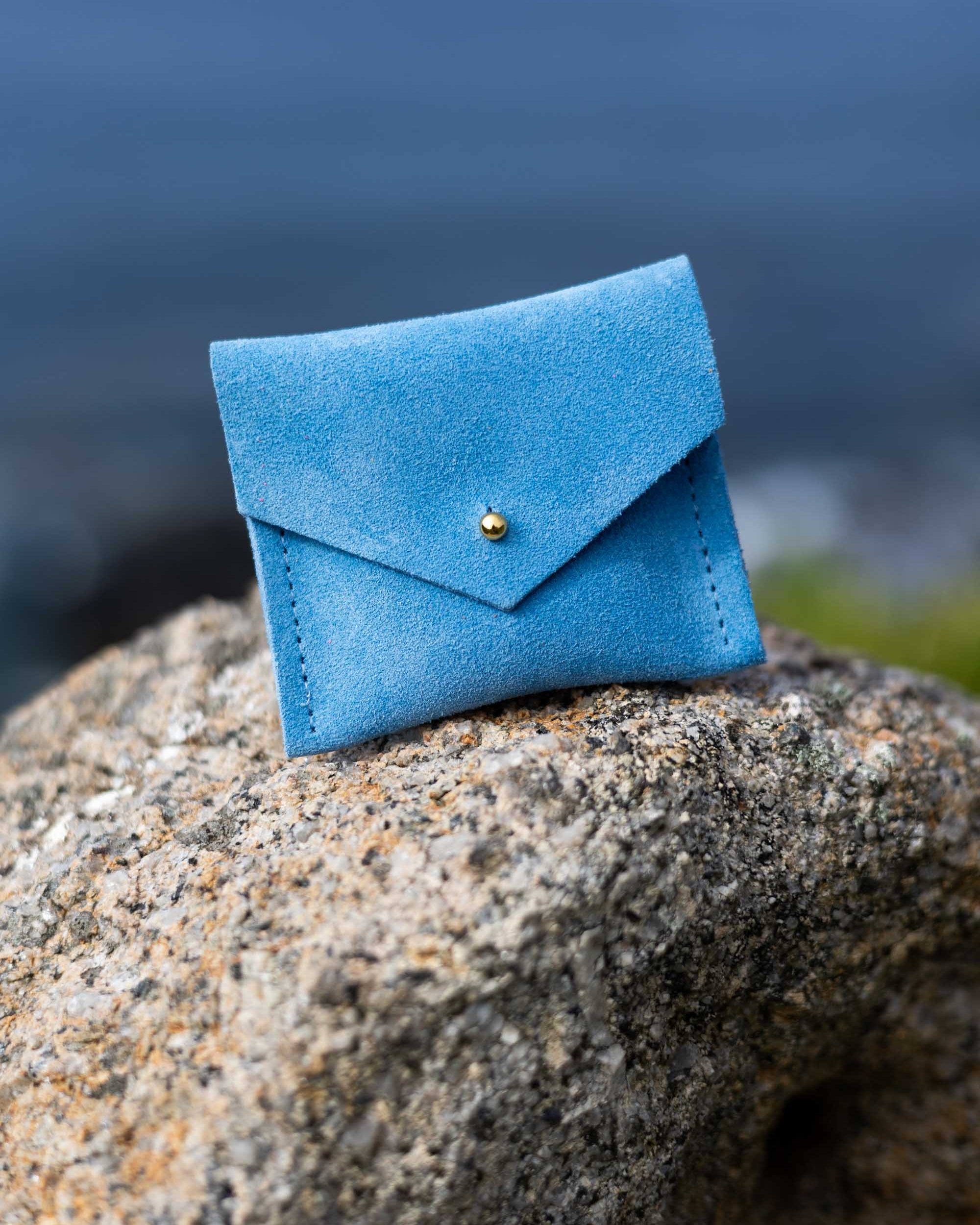 A small blue suede envelope-style pouch with a gold button, resting on a large textured rock with a blurred outdoor background.