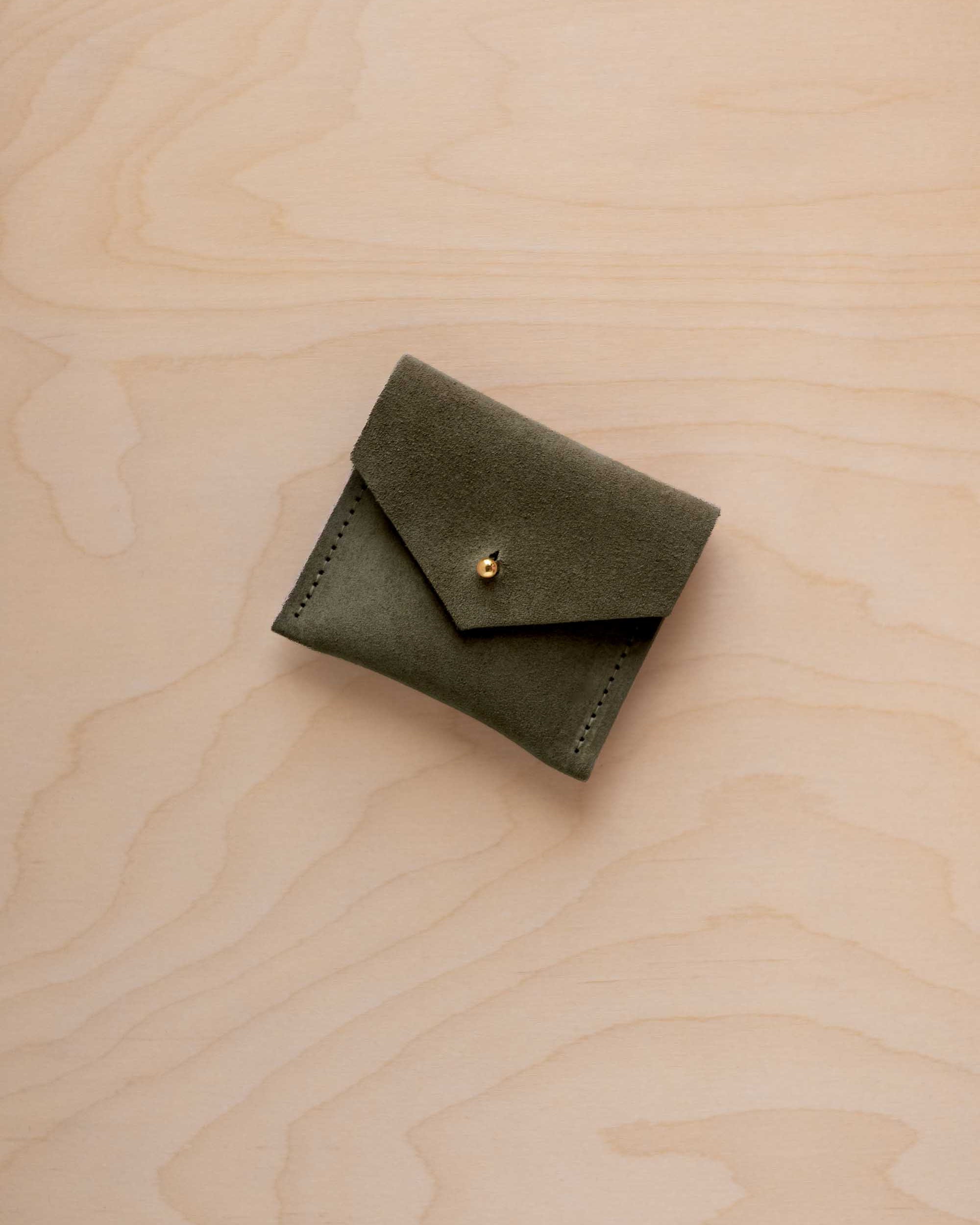 A small olive green suede pouch with a flap closure and gold button, placed on a light wood grain surface.